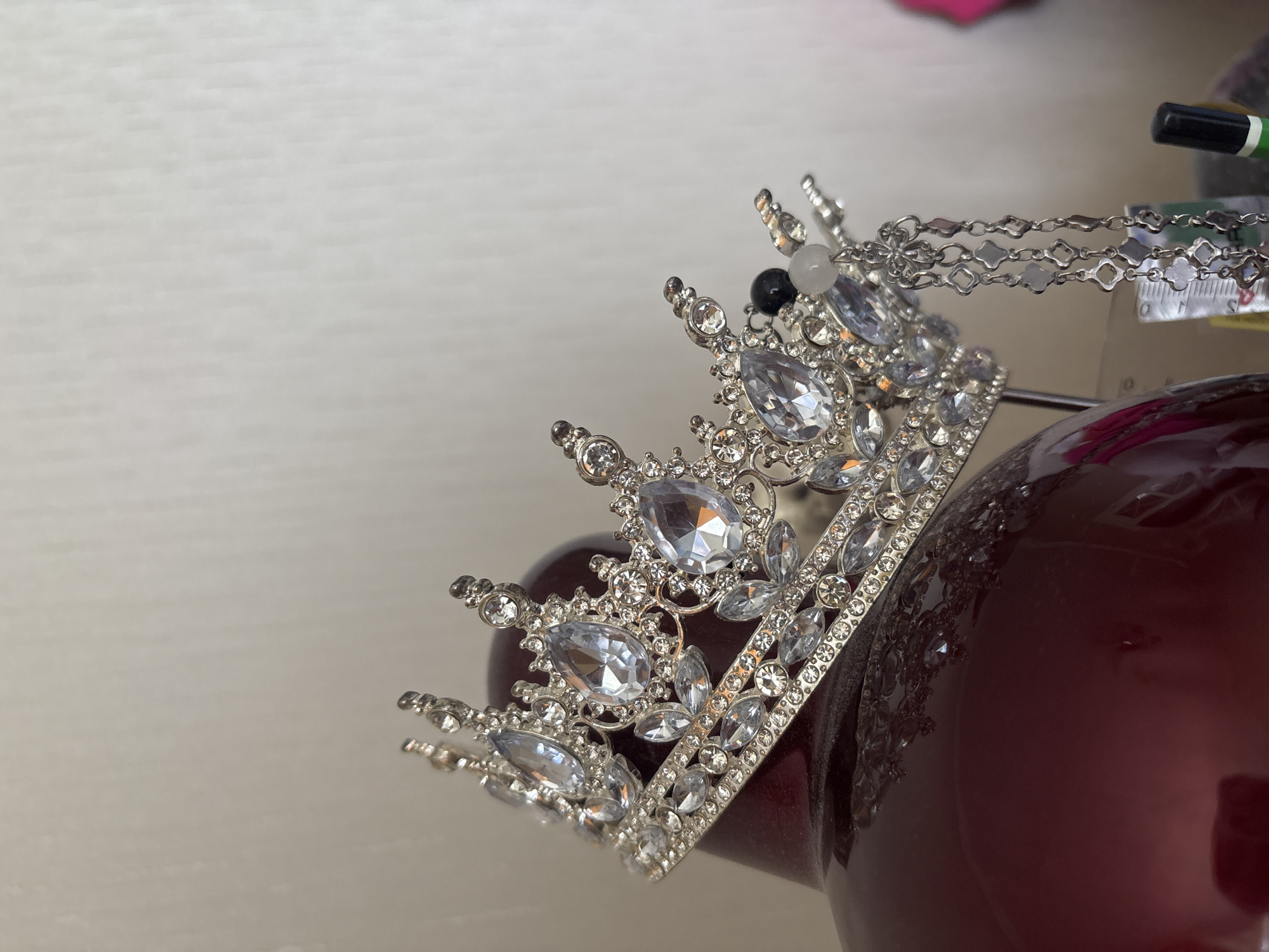 A tiara adorned with clear gemstones rests on a dark red, glossy spherical object. The tiara has a silver-toned metal base with intricate filigree designs and is studded with teardrop-shaped and round clear stones, as well as smaller clear crystals along its band. Dangling from one side of the tiara are several silver chains with small, quatrefoil-shaped metallic charms. A single black bead and a small white bead are also visible attached to the tiara. The background is a plain, light-colored wall with a subtle textured pattern. There are no people or animals in the image, and the scene appears to be indoors. The location in Bila Tserkva, Ukraine, cannot be determined from the visual information provided in the image.