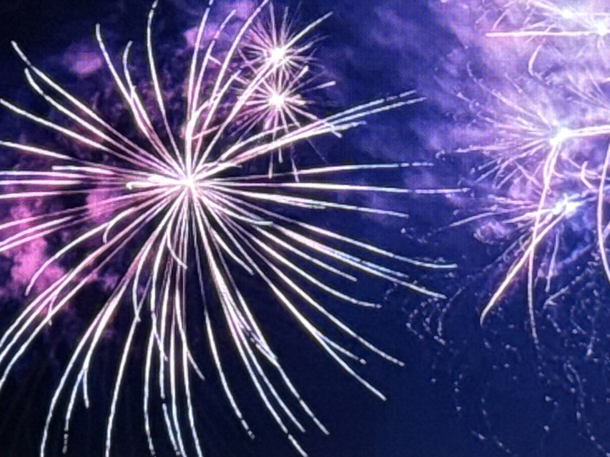 The image depicts a nighttime scene of fireworks exploding against a dark blue sky. The fireworks are primarily white with hints of pink and purple. Several bursts are visible, with radiating lines of light extending outwards from a central point. Some smaller, scattered sparks are also present in the sky. The lower portion of the image shows more fireworks with a similar color palette, appearing to be either further away or a different stage of the display. There are no people, buildings, or discernible landmarks to indicate a specific location within the city of Norilsk, Russia. The focus is solely on the pyrotechnic display in the night sky.
