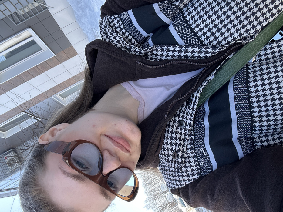 This close-up, daylight outdoor image depicts an individual, likely female, captured in a selfie in Новое Шигалеево, Russia. The subject wears large brown-framed sunglasses, a light pink inner shirt, a dark brown zippered jacket, and an outer vest or jacket featuring a black and white houndstooth pattern with horizontal black and white striped bands. A green strap, likely from a bag, crosses diagonally over the subject's chest. The subject has long, light brown hair, visible behind the left ear, and exhibits a subtle smile. The background reveals a modern multi-story building with a facade clad in alternating light brown/beige and dark grey/silver panels, featuring white-framed windows and an external air conditioning unit. Bare tree branches are visible to the left, and patches of white snow are present on the ground, indicating a cold weather season. The sky is clear.
