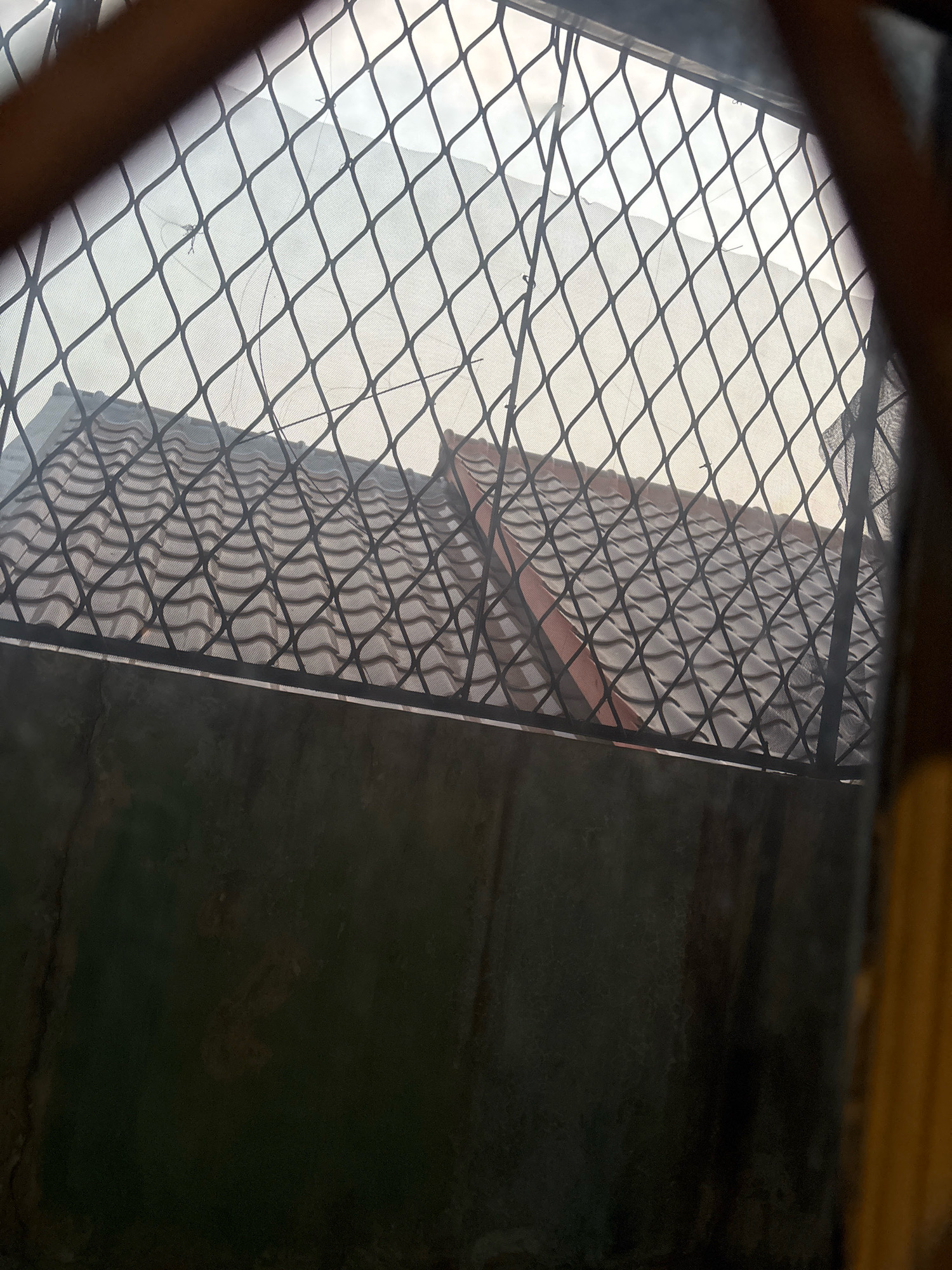 The image shows a view through a black mesh screen, obscuring a scene of rooftops under a hazy sky, likely in Bekasi, Indonesia. The rooftops are covered with curved, grey tiles, some with reddish-brown edges, suggesting a residential or commercial area. The mesh screen creates a diamond pattern overlaying the view. In the foreground, a dark, textured surface, possibly a wall or concrete structure, is visible. The overall atmosphere is overcast and somewhat obscured.