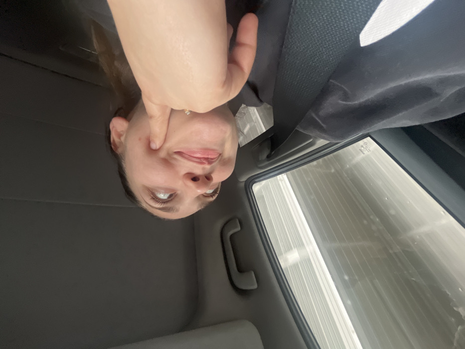 The image depicts the interior of a vehicle, primarily focusing on a female subject positioned in the upper right quadrant of the frame. Her head is tilted, and her eyes are directed upwards and to the left. She is using her right index finger to press into her right cheek, creating a visible indentation and a slight puckering of her lips, indicative of a playful facial expression. A ring with a small, reflective stone is visible on her right ring finger. She is wearing a dark-colored garment. The vehicle's light-colored headliner is visible above and to the left of her head, and a grey overhead grab handle is present in the middle left. A vehicle side window occupies the lower left portion of the image, through which a blurred exterior environment is visible, characterized by light-colored horizontal striations, consistent with a building facade or interior wall. A dark seatbelt strap crosses diagonally from the upper right toward the lower right, partially obscuring the subject's torso. The scene is located in Biss'one, Switzerland.