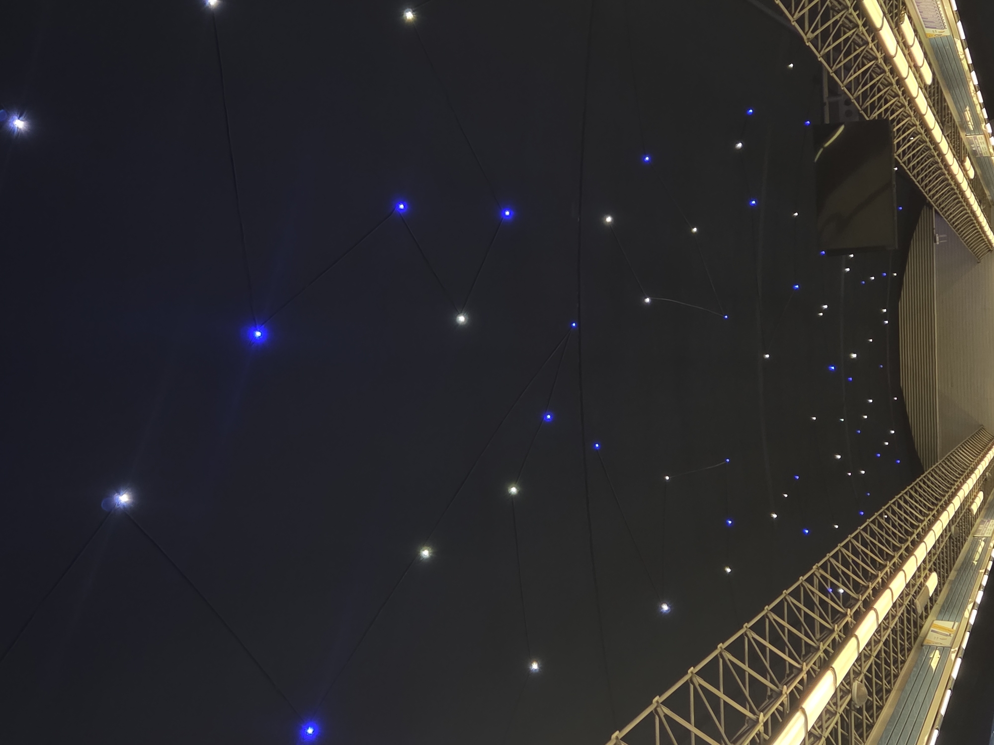 The image appears to be an upward view within an urban environment at night. A series of lights, alternating between white and blue, are strung up against a dark background, perhaps a ceiling or open sky. The lights are connected by dark lines, giving the impression of constellations. On the right, there is a structural element, possibly part of a building or bridge, constructed with a lattice or grid-like pattern. This element is illuminated, casting a warm, artificial glow. The angle suggests the photo was taken looking straight up from below.