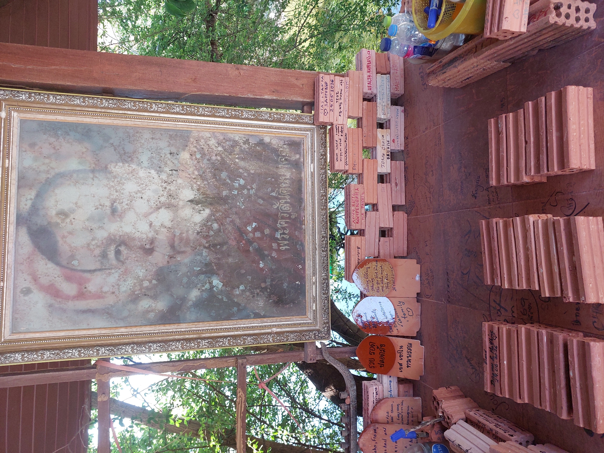 The image depicts an outdoor scene at Baan Plai Laem, Thailand, likely a temple or memorial site. A large, faded portrait in a golden frame hangs prominently, showing the upper body of an elderly man with indistinct features. Below the portrait, several rows of un-fired terracotta bricks are stacked. Many of these bricks have been inscribed with names, dates, and messages in various languages, including Thai and English. The ground surface, also covered in inscriptions, appears to be a wooden platform or paved area. Several water bottles and a yellow container with blue markers are visible to the right of the stacked bricks. In the background, lush green foliage and trees suggest a natural, possibly tropical, environment. The overall impression is of a place where visitors leave personal dedications or wishes.