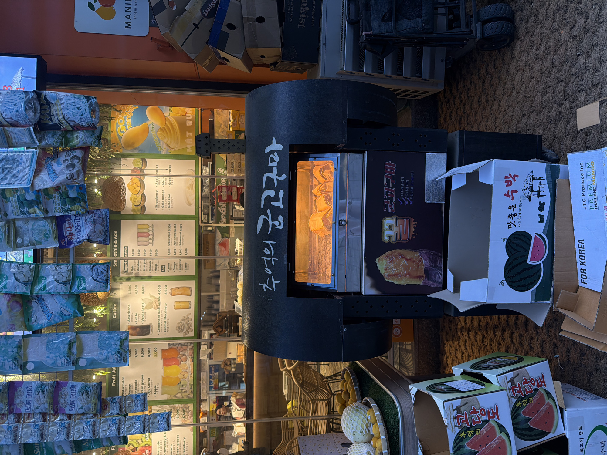 The image shows an outdoor food stall in Seoul, South Korea. A black cylindrical oven with "추억의 군고구마" (Memories of Roasted Sweet Potatoes) written in Korean on its side is the focal point. Inside the oven, sweet potatoes are being roasted. To the right of the oven are several open cardboard boxes with watermelon prints on them and the words "맛좋은 수박" (Delicious Watermelon) and "FOR KOREA." One box also displays "JTC Produce Inc. PREMIUM THAILAND MANGO." Behind the oven, a glass window displays a menu with items like fruit juice, coffee, and smoothies along with their prices. Posters of mango smoothies are also visible. To the left of the menu is a display of various snack packages. Baskets containing fruits are visible at the lower edge of the image, along with chairs. Above the stall, there are stacked boxes with labels. The ground is textured and appears to be a paved surface. The image suggests a vibrant street food scene with various Korean and imported products.