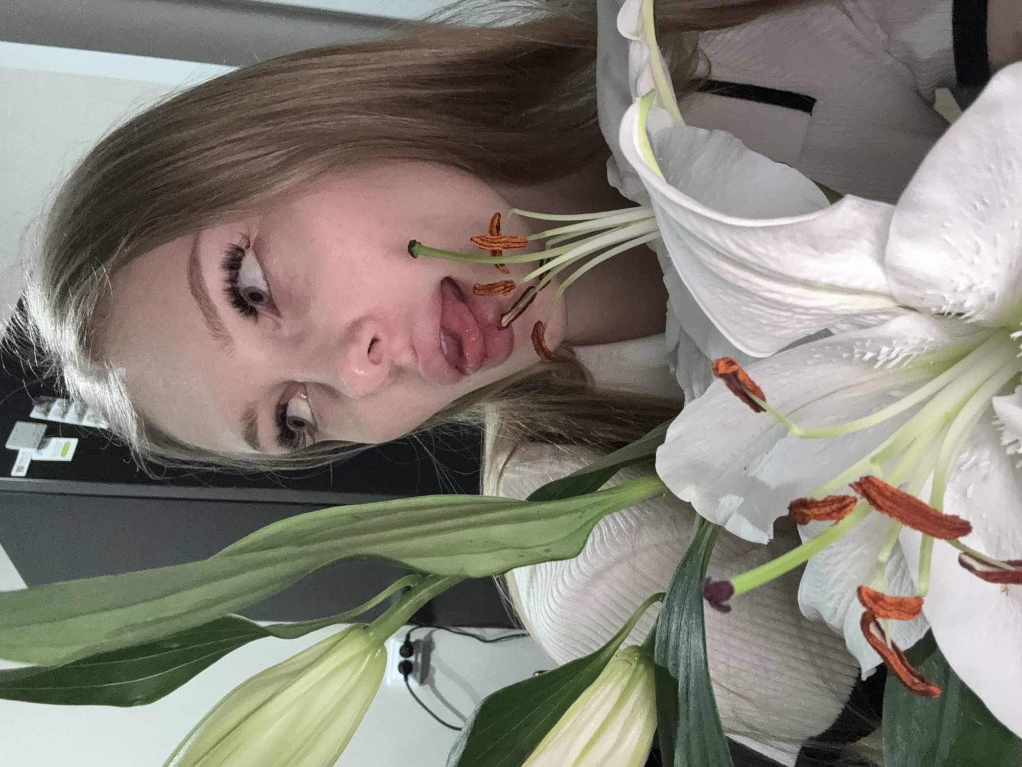 A young woman is positioned behind a bouquet of white lilies. Her face is angled towards the camera, with her tongue extended. The flowers are in full bloom, with prominent stamen and green leaves. The background shows a glimpse of a computer keyboard and a light-colored wall with electrical outlets. There is no indication of the location as the city of Perm, Russia.