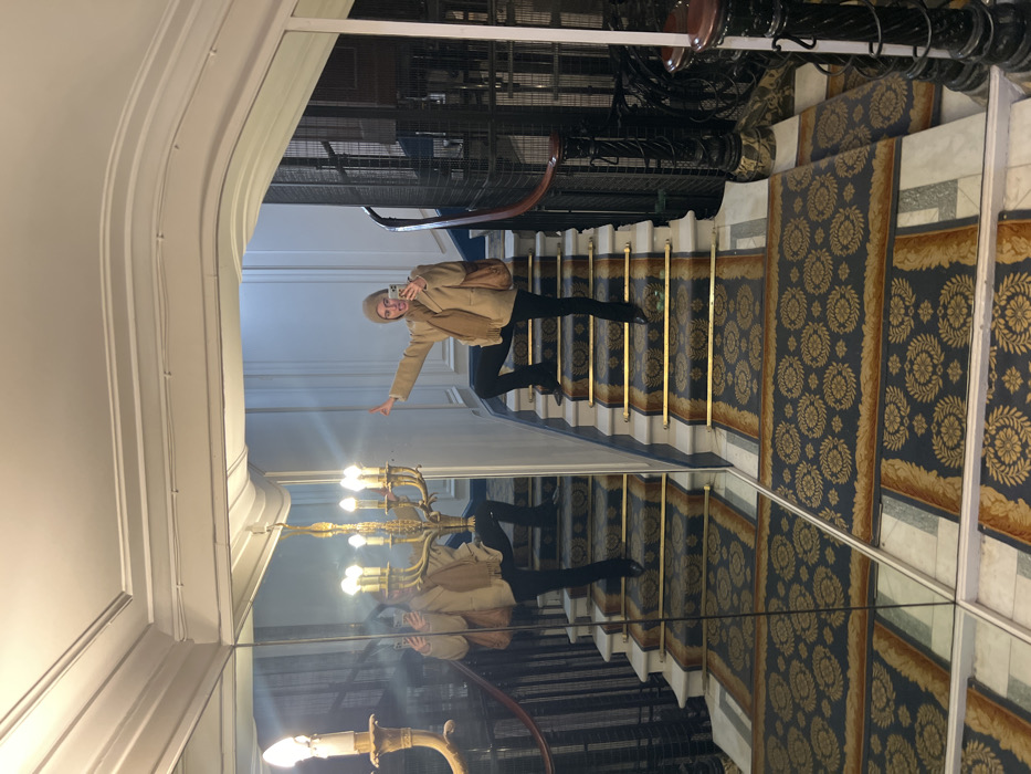 A person stands on a staircase with a blue and gold patterned carpet. They wear a beige coat, black pants, and a beige hat, holding up a phone for a photo. The staircase has gold rails. The left side of the image is a mirror, reflecting the person and the stairs. Above, a vintage metal elevator structure is visible. The walls are white with decorative molding. A gold chandelier hangs from the ceiling near the mirror.