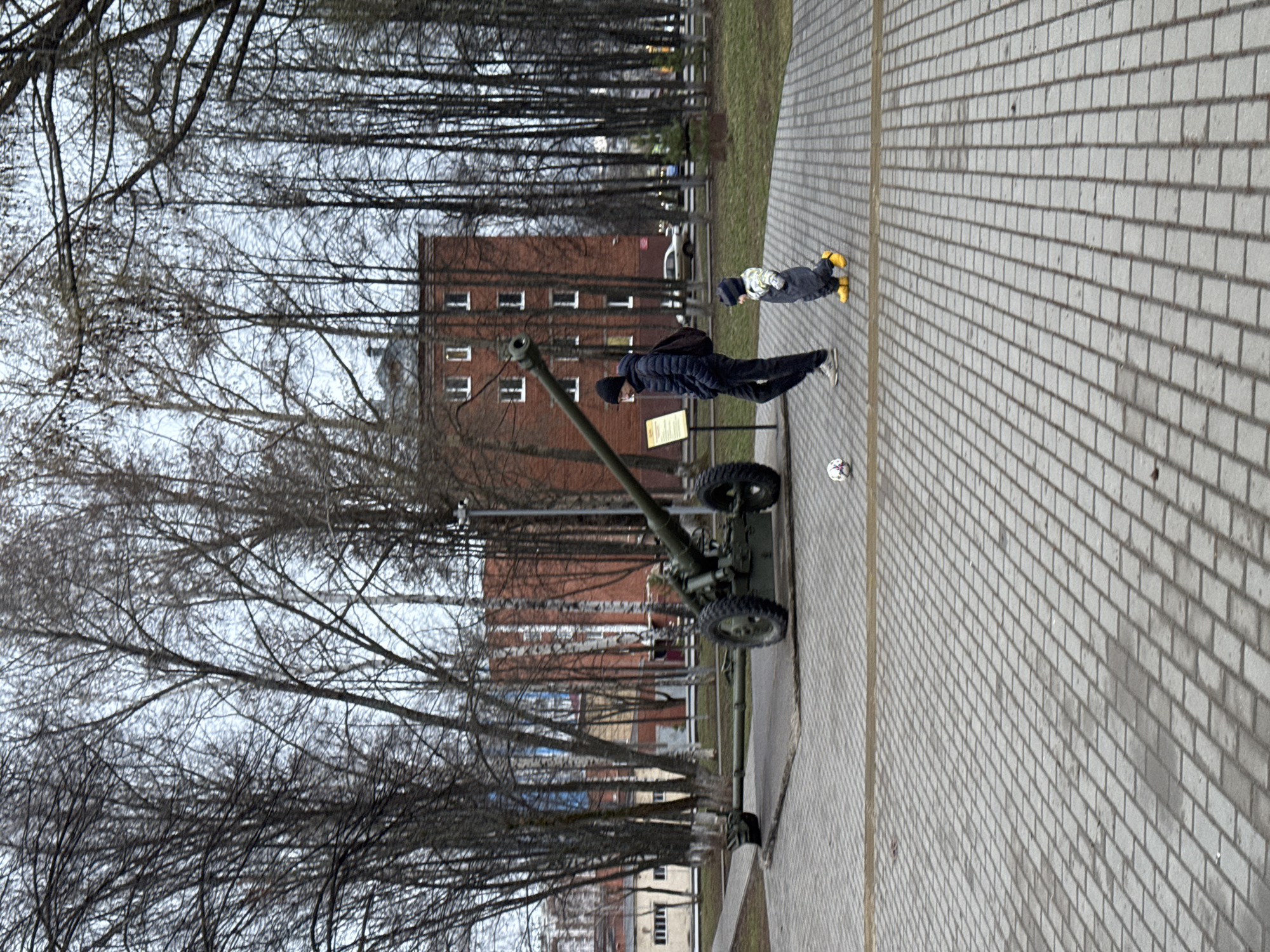 The image shows an outdoor scene in Izhevsk, Russia. A dark green cannon, likely a war memorial, stands on a paved walkway. An adult male with a dark jacket and black hat stands near the cannon. A small child is a few feet away, wearing a dark and light patterned jacket and yellow boots. A white soccer ball is near the child. The brick-paved walkway extends into the distance. Bare trees are visible, lining the background. Several brick buildings are in the background.