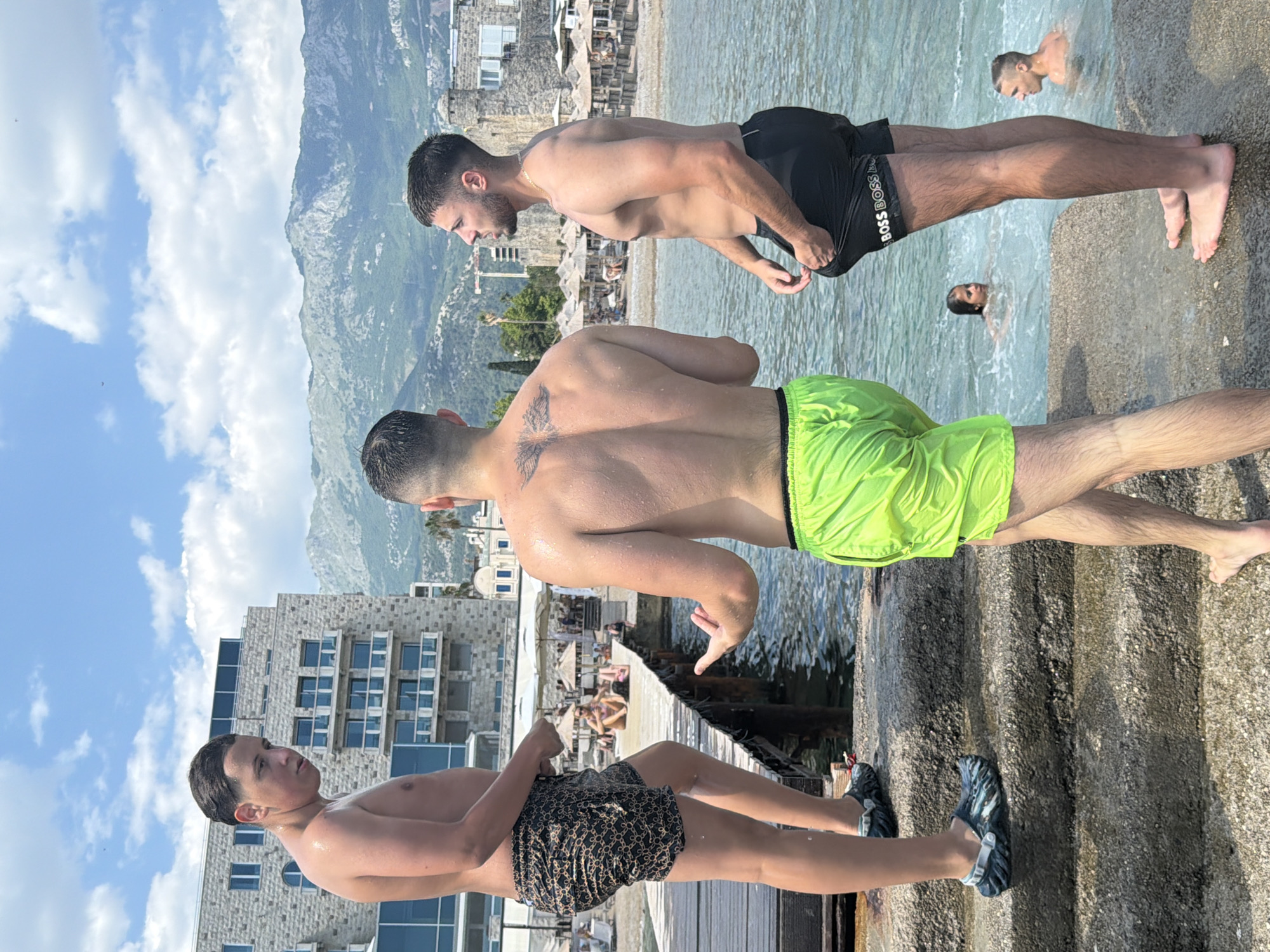 Three young men are standing on a stone staircase beside the Adriatic Sea in Budva, Montenegro. The man on the left is facing away from the camera, looking towards the water. He is wearing patterned swim shorts and is standing on a wooden pier. The man in the center is facing the man on the right, with his back to the camera. He has a tattoo on his back and is wearing bright green swim shorts. The man on the right is facing away from the camera and looking towards the sea. He is wearing black swim shorts with "BOSS" printed on them. In the water behind them, two people are swimming. Further back, there is a beach with umbrellas and sunbathers, and a large stone building and mountains are visible in the background. The sky is blue with scattered clouds.