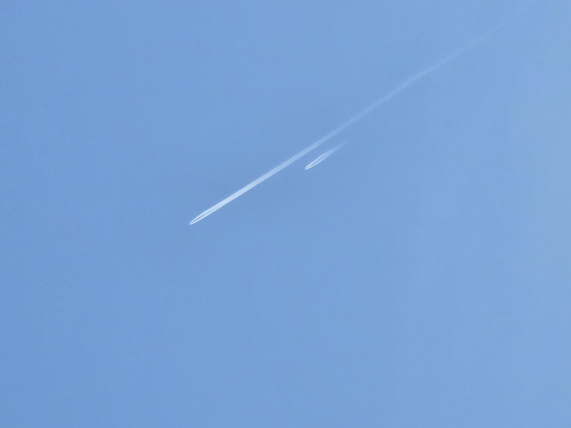 The image shows a clear blue sky with contrails from two airplanes. One airplane is in the foreground, leaving a long, distinct contrail, while another airplane is slightly behind it and leaving a shorter, less distinct contrail. The location is not discernible from the image.
