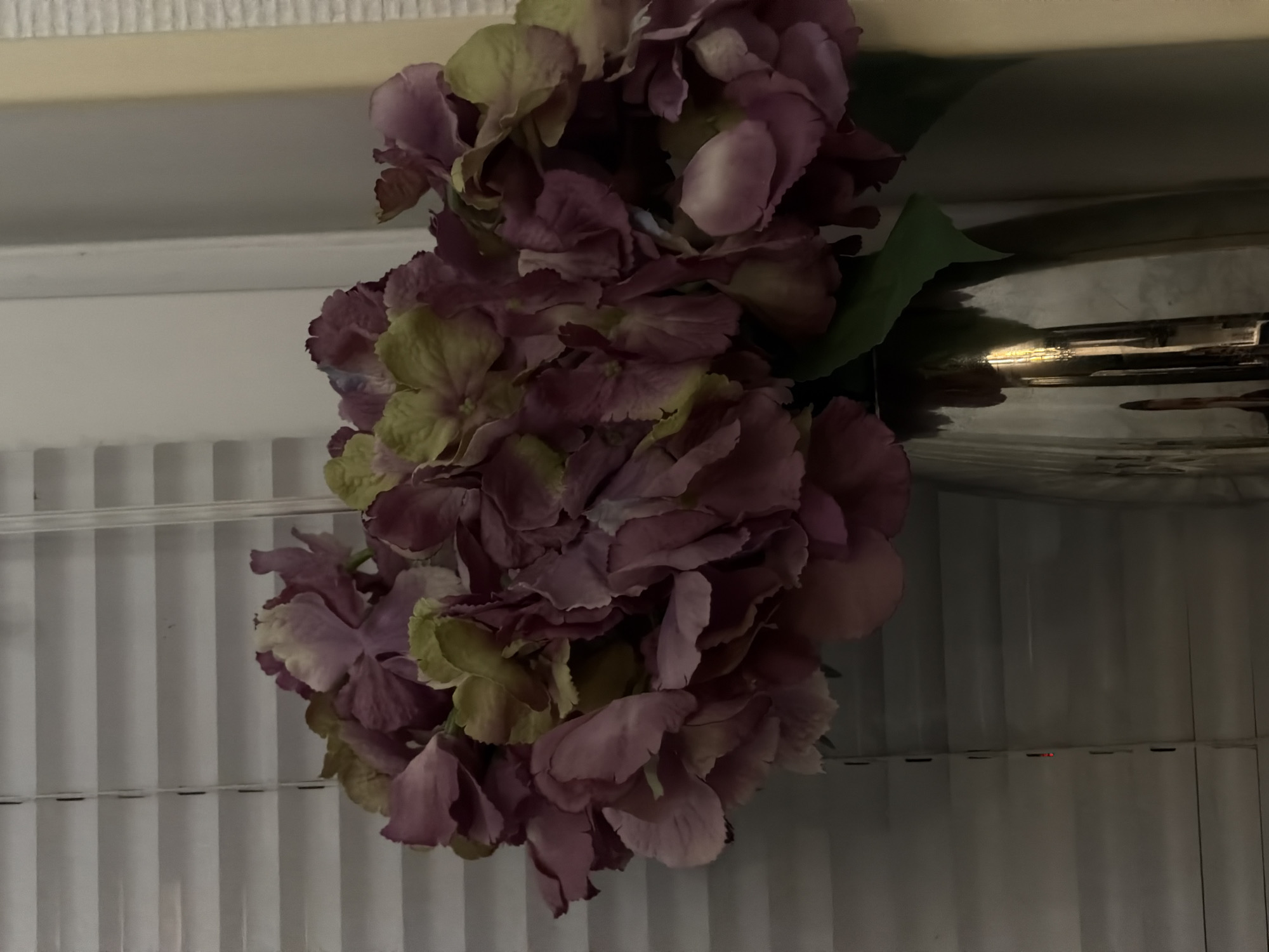 A close-up, indoor shot shows a bouquet of artificial hydrangeas in shades of purple and green, arranged in a shiny silver vase. The vase sits in front of closed white blinds. The lighting is dim, with a warm glow suggesting artificial illumination. The background to the right of the vase is a neutral, light-colored wall or curtain. There are no people or animals visible in the image. The scene appears to be a still life arrangement in a domestic setting, possibly a home or an office, in Bila Tserkva, Ukraine.