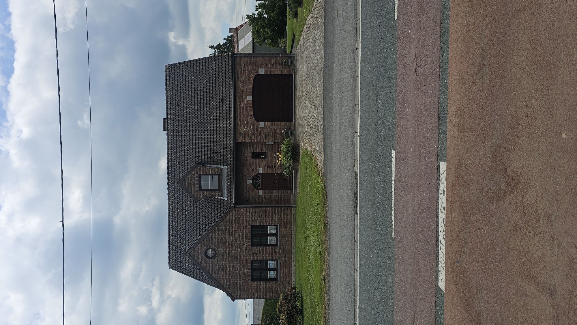 The image shows a brick house with a dark tiled roof, featuring a prominent arched garage door. There is a small lawn in front of the house, bordered by a paved driveway. A street with pedestrian crossing markings is visible in the foreground. The sky is overcast with clouds. The house is situated in Boussu-lez-Walcourt, Belgium, indicated by external data. No people or animals are visible in the scene.