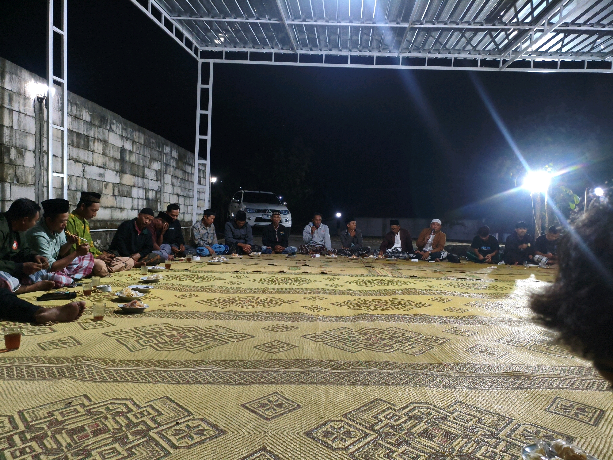 An outdoor gathering of approximately 15-20 men is taking place at night, seated on a large, woven yellow mat on the ground. The mat has a repeating geometric pattern. In the background, a white brick wall is visible, and beyond that, a white SUV is parked. A white metal structure with a transparent roof covers the area where the men are seated. The lighting suggests it's a nighttime event, with bright lights creating lens flares. The men are dressed casually, some wearing traditional Islamic caps. Several small plates with food and glasses of drinks are scattered on the mat in front of the men. The overall atmosphere appears to be informal and communal, possibly a religious or social gathering. The location is identified as Segiri, Indonesia.