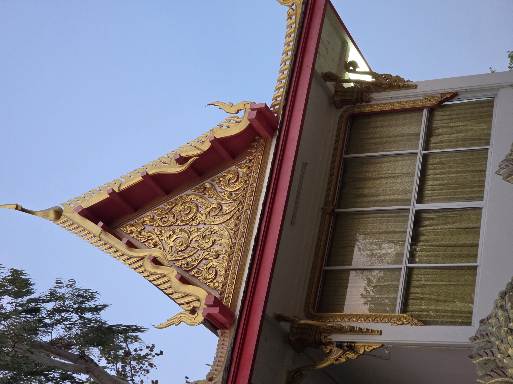 The image captures the architectural details of a building, likely a temple, in Pattaya City, Thailand. The structure features ornate gold carvings on a triangular roof section with a red border. Adjacent to the roof is a window with a white frame and gold decorative elements. The sky is light blue. The bottom left of the frame includes a tree.