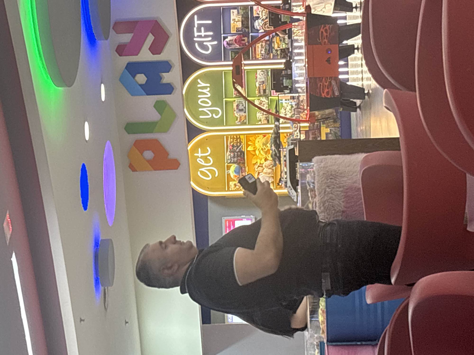 The image depicts an indoor scene within what appears to be a modern commercial or entertainment establishment, likely located in Yerevan, Armenia. The primary subject is a man, shown from the chest up, standing in the mid-ground. He wears a black polo shirt with white trim on the collar and sleeves, paired with dark trousers, and holds a dark mobile device in his right hand. He is looking slightly upwards and to the right.
Behind the man, a prominent wall display features large, colorful, block-style letters spelling "PLAY" in shades of pink, blue, green, and orange. Below this, there are three illuminated, arched display shelves, each with a different colored background and text: "get" (yellow), "your" (green), and "GIFT" (blue). These shelves showcase various items, possibly toys or gifts.
To the right of the man, several red, contemporary-designed chairs with high, curved backs are visible, suggesting a seating or waiting area. A fluffy white cushion rests on a surface near these chairs. The upper wall and ceiling feature colorful, illuminated circular and oval-shaped lights in green, blue, and purple. In the background, a red and black arcade-style machine or display with distinctive patterns can be partially seen. The overall environment is brightly lit and modern.