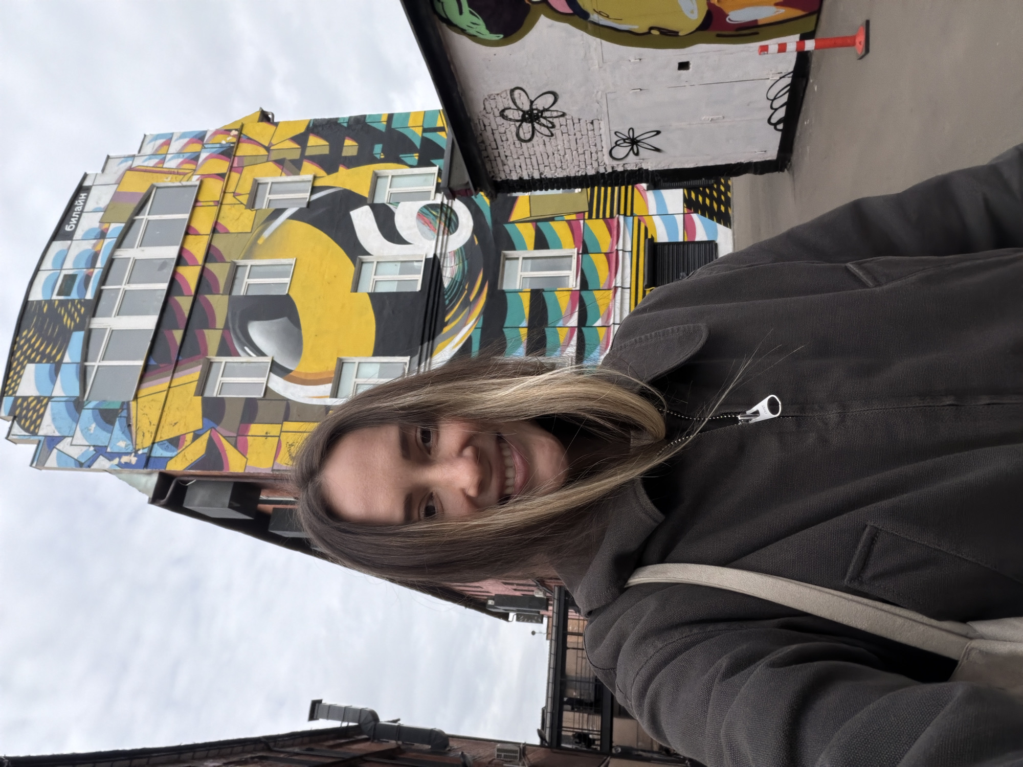 A female subject, smiling directly at the viewer, occupies the foreground of an outdoor urban scene. She has long, straight brown hair with blonde highlights and wears a dark gray zippered jacket, with a light-colored strap of a bag visible over her right shoulder. In the background, a multi-story building in Khimki, Russia, is prominently featured, extensively covered in a colorful abstract mural. The mural incorporates yellow, black, blue, and white geometric patterns, including a large stylized number '6'. The word "БИЛАЙН" (Beeline) is visible in black Cyrillic letters at the top of this building. To the right of the subject, a lower white brick wall displays black line-art graffiti of flowers. An orange and white striped traffic cone is on the gray paved ground nearby. To the left of the subject, a reddish-brown brick building with exposed dark piping and a multi-level structure with a glass walkway are partially visible. The sky is overcast.