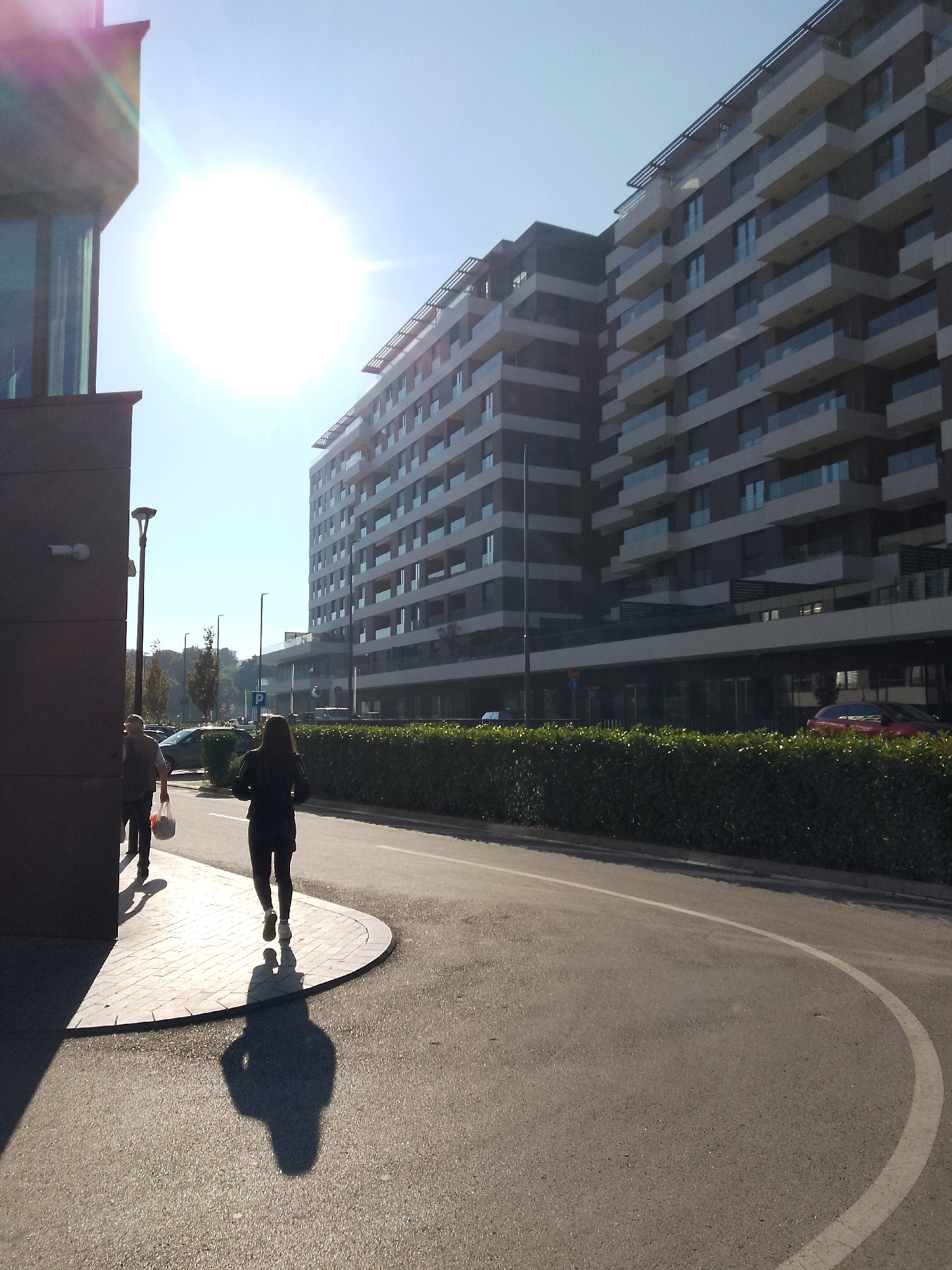 This is a vertical, outdoor, daytime photograph taken at an eye-level perspective, showing a scene in Belgrade, Serbia. In the foreground, a woman is walking away from the camera on a paved sidewalk with a curved edge. She is wearing dark clothing, including leggings and a jacket, and white sneakers. Her shadow is cast prominently on the sidewalk and the adjacent asphalt road. To the left of the woman, a man is also walking away from the camera, carrying a white plastic bag. He is wearing a dark backpack.
Behind the people is a modern, multi-story building with a dark facade and white balconies. Further in the background, another tall building is visible, also featuring multiple balconies with glass railings. The buildings are set against a clear, bright blue sky. A bright sun is positioned in the upper left quadrant of the image, causing lens flare and overexposing the sky in its vicinity.
A well-maintained green hedge runs along the edge of the road, separating it from the buildings. Cars are parked in the background. A street lamp is visible to the left of the woman. The overall environment suggests an urban setting with contemporary architecture and sunny weather.