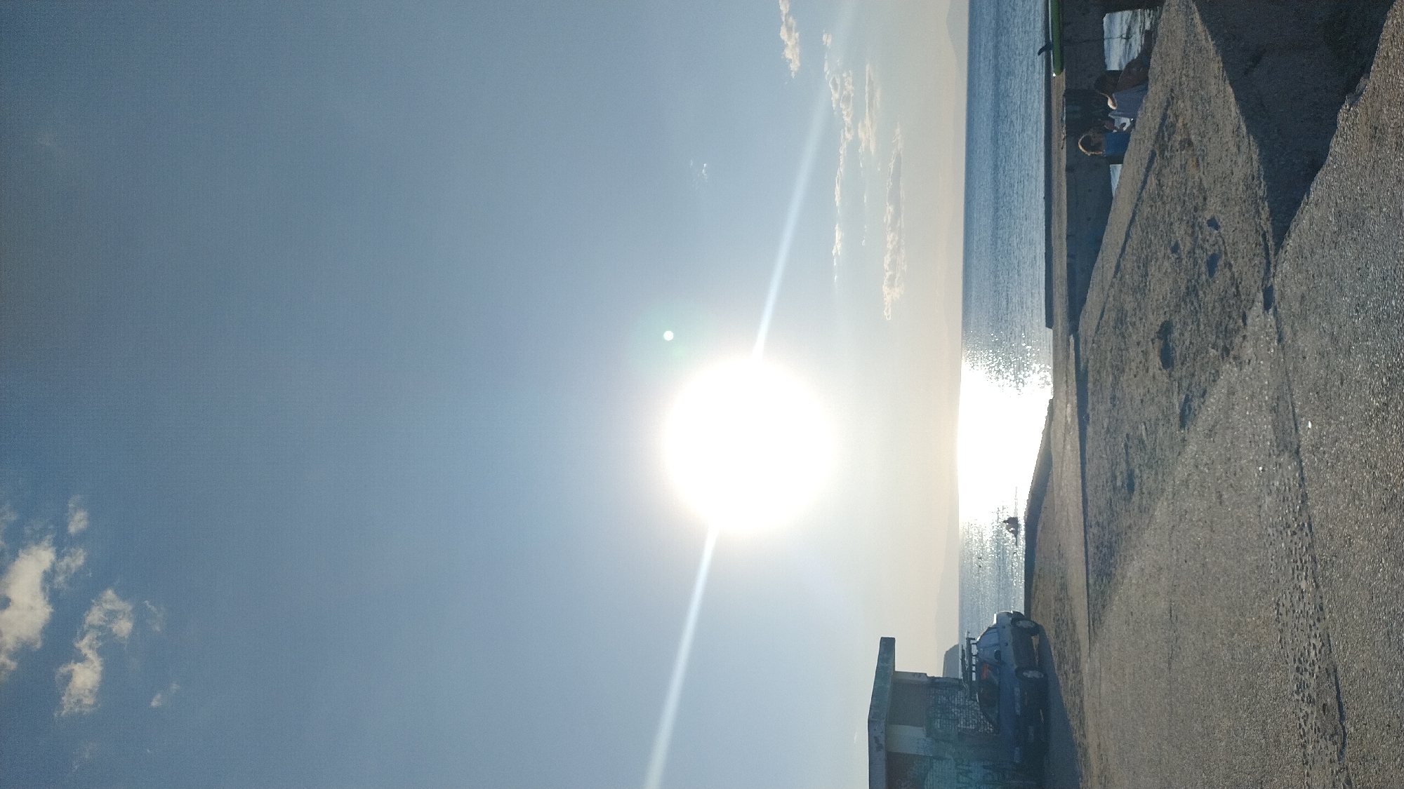 The image depicts a coastal scene in Perea, Greece, bathed in the bright, low sunlight of either sunrise or sunset. The sun is prominently centered in the frame, casting a strong glare and reflecting a shimmering path across the calm, blue sea. In the foreground, a concrete paved area, possibly a harbor or promenade, slopes gently towards the water. To the left, a small, weathered, multi-story building stands with graffiti on its lower section and a car with a roof rack parked beside it. Further along the shoreline, a few individuals are visible near the water's edge, and some appear to be sitting on a concrete structure to the right, possibly a jetty. The sky is a clear blue with a few wispy clouds, and distant landmasses are faintly visible on the horizon.