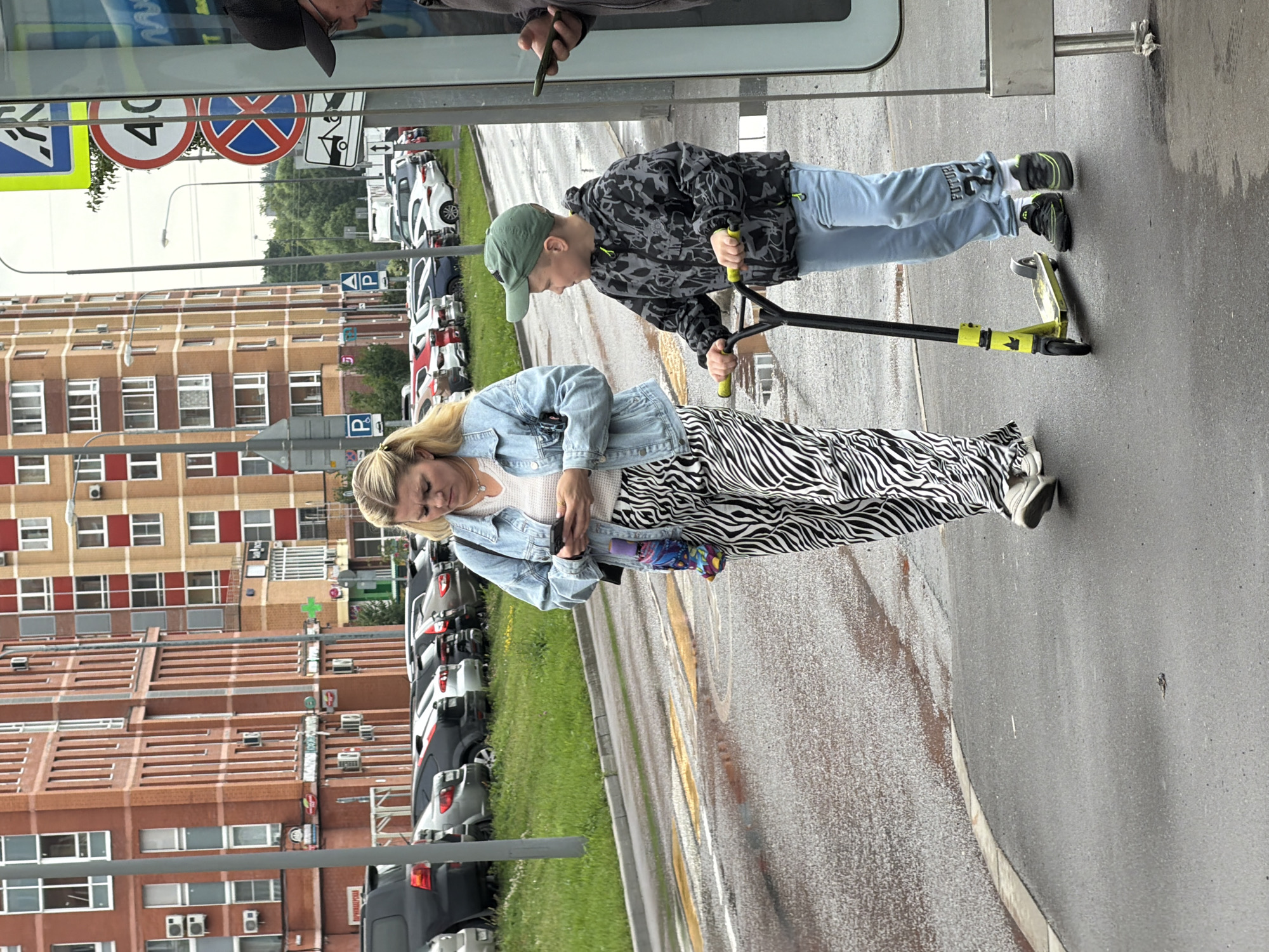 In Shcherbinka, Russia, a woman and a young boy are walking on a wet asphalt path. The woman, wearing a denim jacket and zebra-striped pants, is looking at her phone. She carries a colorful umbrella. The boy, dressed in a camouflage jacket and light blue pants, rides a yellow and black scooter. He is wearing a green baseball cap and black sneakers. In the background, several cars are parked in front of a brick building. Traffic signs, including a "40" speed limit sign and a no-parking sign, are visible. The environment suggests it is a cloudy day, possibly after rain.