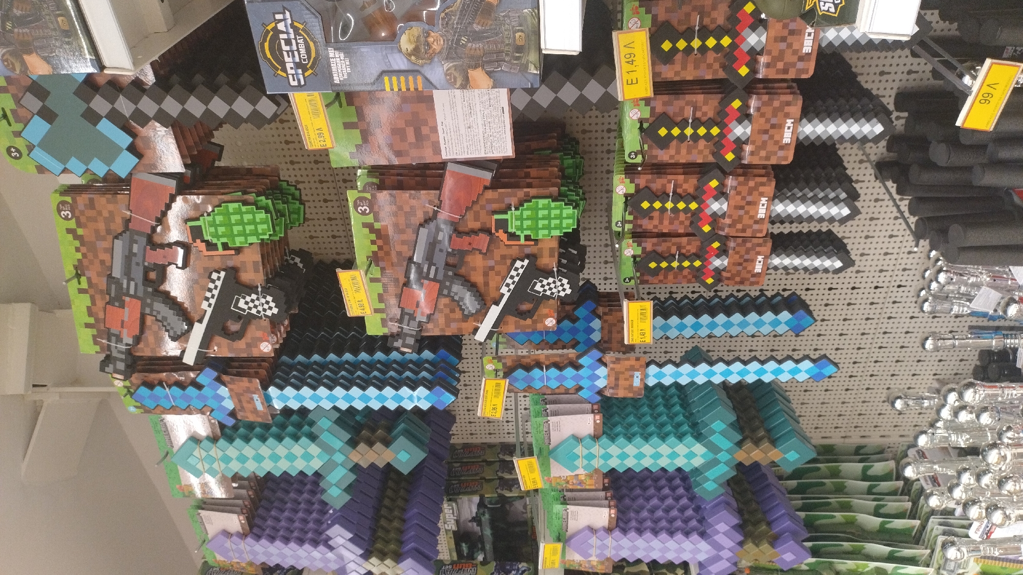 The image shows a retail display of Minecraft-themed toy weapons in a store. The items are primarily pixelated swords, axes, and guns, made of foam or plastic, packaged on cardboard backing designed to resemble the game's blocky aesthetic. Several price tags are visible, indicating prices in Euros, with one reading "E 4,99 л." and another "E 1,49 л." The display is arranged vertically on a pegboard wall. In the foreground, there are rows of other toys, including what appear to be silver-tipped staffs and dark, cylindrical objects, possibly foam batons or handles. The lighting suggests an indoor retail environment. The specific location is a store in Pylaia Municipal Unit, Greece.