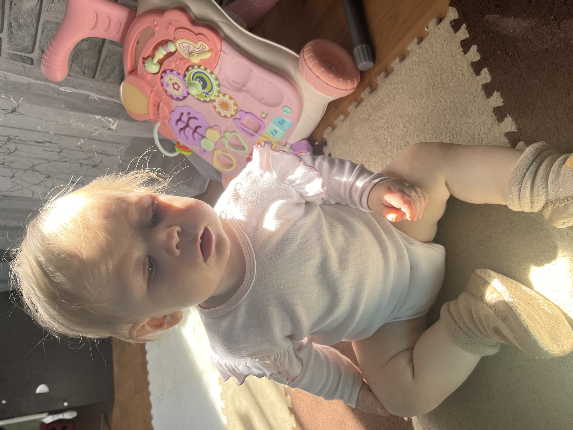 A light-skinned baby with blonde hair is depicted sitting on the floor indoors in Vesyoloye, Russia. The baby wears a light pink, long-sleeved onesie with ruffled detailing on the sleeves and light-colored socks or soft shoes. The baby's head is tilted back, with a slightly open mouth, looking upward.
Beside the baby is a pink baby walker or activity center. This toy features various colorful interactive elements, including gears, shapes, and buttons. The text "Fa So" is visible on one of the toy's buttons.
The floor beneath the baby and toy is covered with interlocking puzzle mats, alternating between beige and brown sections. Strong, natural sunlight streams into the room, casting bright highlights on the baby's hair and clothing, as well as on parts of the floor and the toy, indicating a morning or late afternoon time of day. A dark, cylindrical object and other background elements are faintly visible beyond the immediate play area.