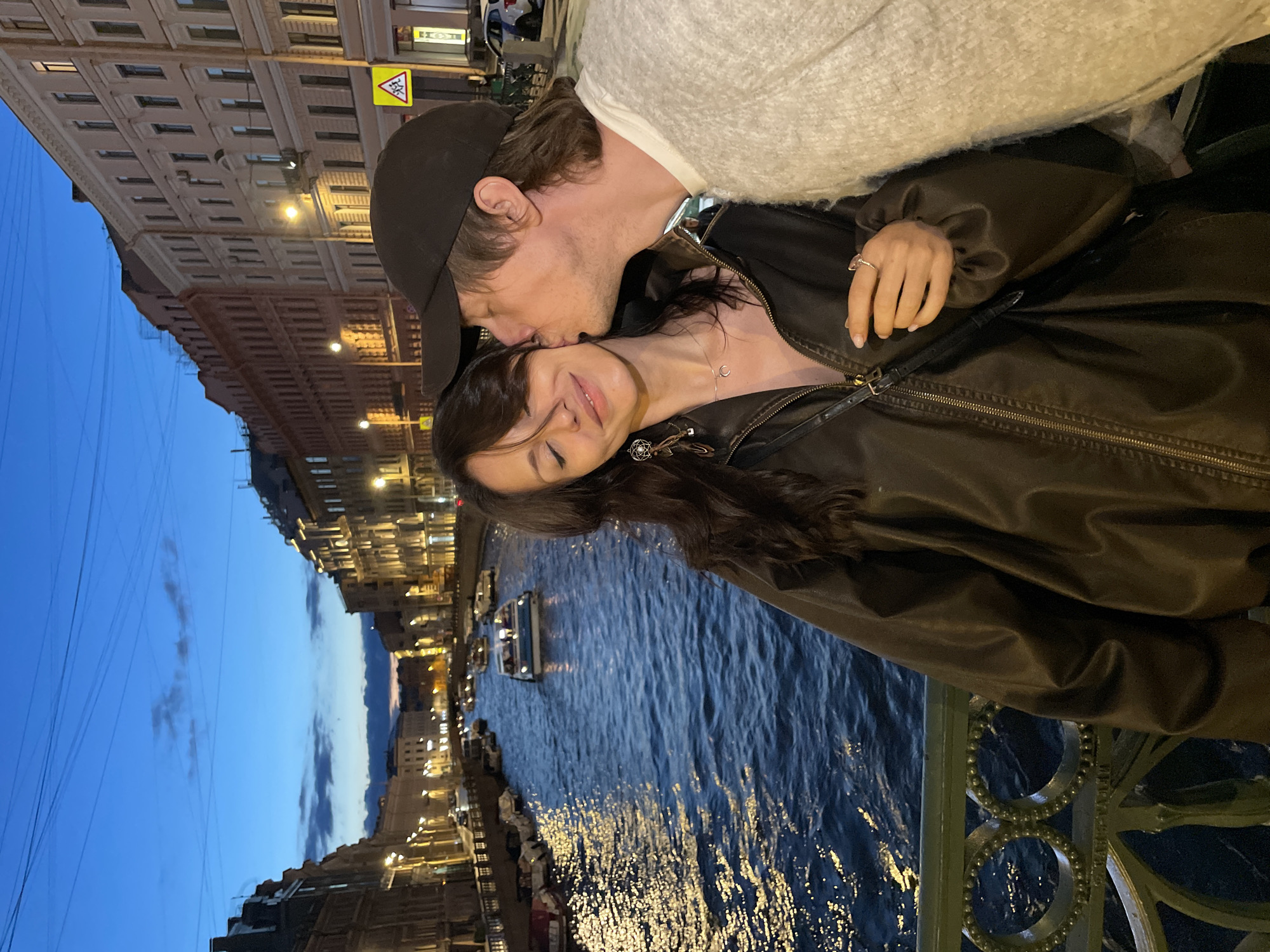 The image captures a romantic moment between a couple at dusk along the Griboyedov Canal in Saint Petersburg, Russia. The man, wearing a dark baseball cap and a textured off-white sweater, is gently kissing the woman on the cheek. She has her eyes closed and a serene expression, wearing a dark brown leather jacket, a delicate silver necklace, and distinctive earrings. A black crossbody bag is slung over her shoulder. They are standing on a decorative green railing overlooking the canal. The canal is filled with dark blue water, reflecting the warm lights from the buildings and street lamps lining its banks. Several boats are visible on the water, some docked and others cruising. The architecture of the buildings flanking the canal is characteristic of Saint Petersburg, with multi-story facades and illuminated windows, creating a picturesque urban scene as the sky transitions from twilight blue to a deeper evening hue. A yellow traffic sign with a pedestrian symbol is visible on the right side of the image.