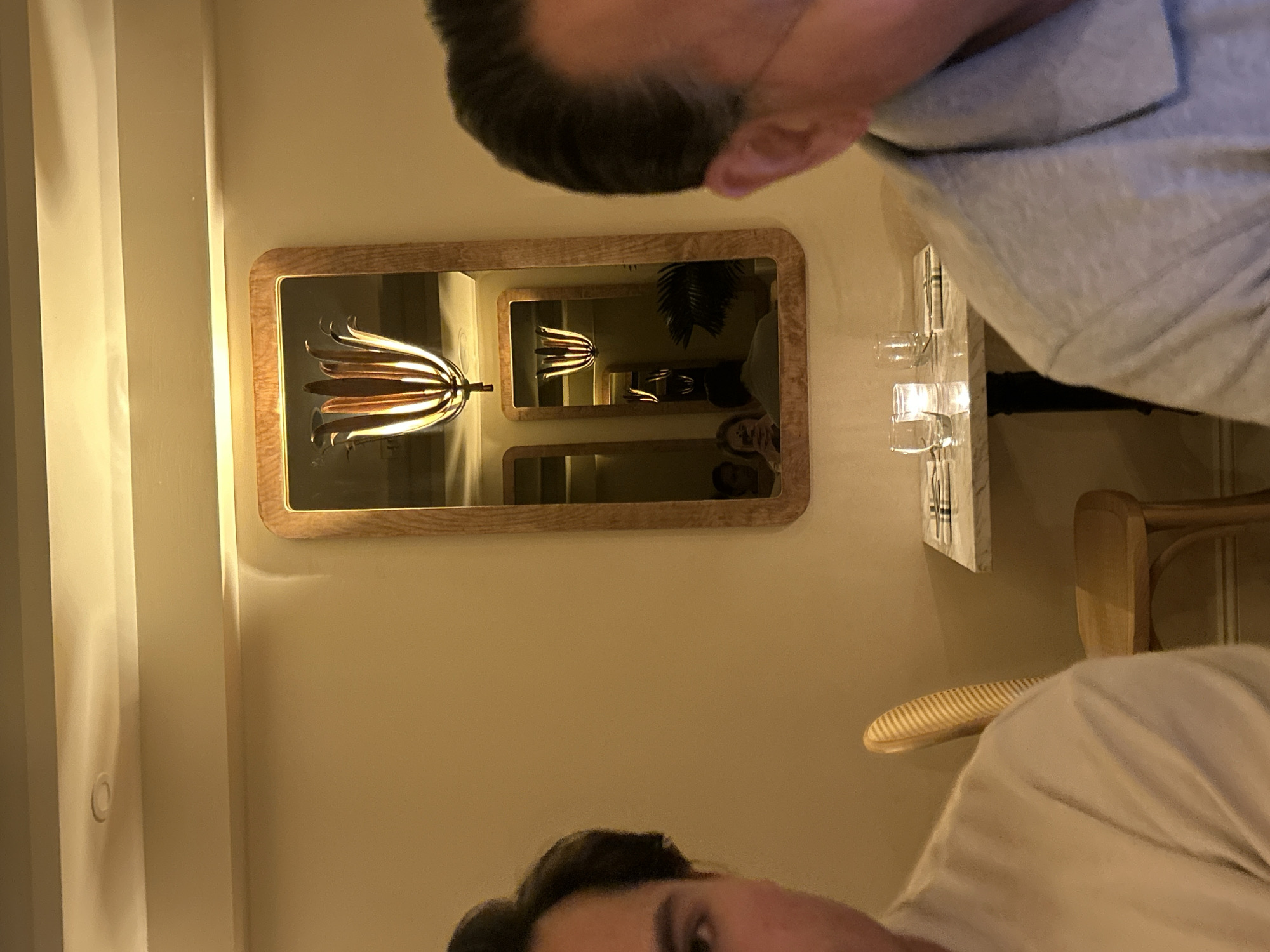The image captures an indoor scene, likely a restaurant or a stylish dining area, illuminated by warm, ambient lighting. A prominent feature is a rectangular mirror with a wooden frame, placed on a light-colored wall. This mirror reflects a distinctive light fixture resembling a cluster of metallic petals or leaves, creating an interesting visual focal point. The reflection includes an "infinity mirror" effect, as a smaller mirror within the larger one reflects the light fixture again.
At least two individuals are visible. In the upper right, the top of a person's head and shoulder (wearing a grey shirt) are seen, looking downwards. In the lower left, another person's forehead and eyes (wearing a lighter shirt) are visible, looking slightly upwards. A third person's face is partially reflected in the main mirror, looking towards the right.
On the right side of the image, a white table surface holds several clear glass items, possibly votive candle holders, which emit a soft light. Part of a wooden chair with a patterned back is also visible. The warm, artificial lighting suggests an evening setting. The overall mood appears relaxed and intimate, consistent with a social gathering or dinner. No visible text is present in the image.