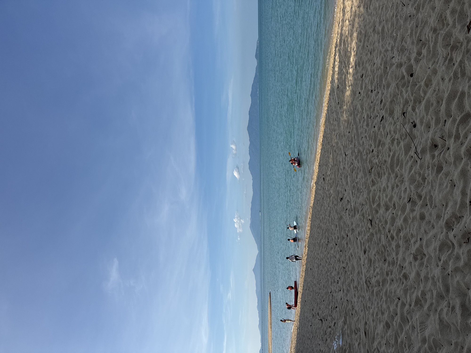 The image depicts a wide, sandy beach on a sunny day in Ko Samui District, Thailand. The foreground is dominated by textured sand, with scattered debris. The midground features a calm, turquoise sea with several individuals engaged in water activities. On the left, a person stands near a red kayak on the shore. Further into the water, a group of people are visible: some standing, some on paddleboards, and a pair in a kayak. In the distance, a chain of lush, mountainous islands forms the horizon under a clear blue sky with scattered white clouds. The overall scene suggests a recreational day at the beach with various water sports being enjoyed.