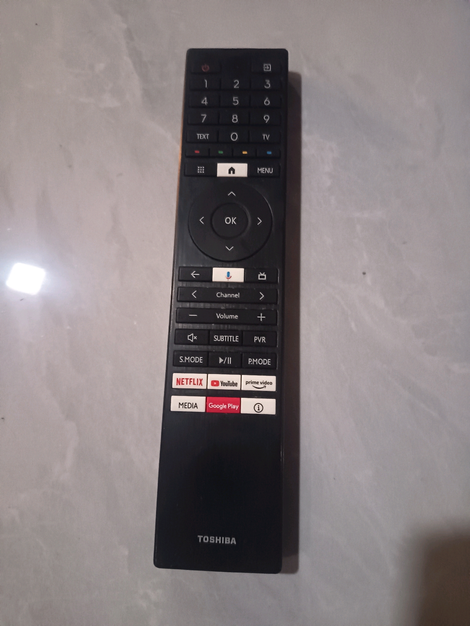 The image shows a Toshiba remote control lying on a light-colored, textured surface, likely a tiled floor. The remote is black with various buttons, including a numerical keypad, navigation controls (arrow keys and OK button), dedicated buttons for Netflix, YouTube, and Google Play, and other function buttons such as Volume, Subtitle, and Menu. The Toshiba logo is visible at the bottom of the remote. The background is a mottled, greyish-white texture that suggests a floor or wall. There are no people or animals present in the image. The lighting in the image is uneven, with a bright glare on the left side, possibly from an overhead light source. The remote is positioned vertically in the center of the frame. The location of Sidorejo, Indonesia is not depicted or indicated in the image.