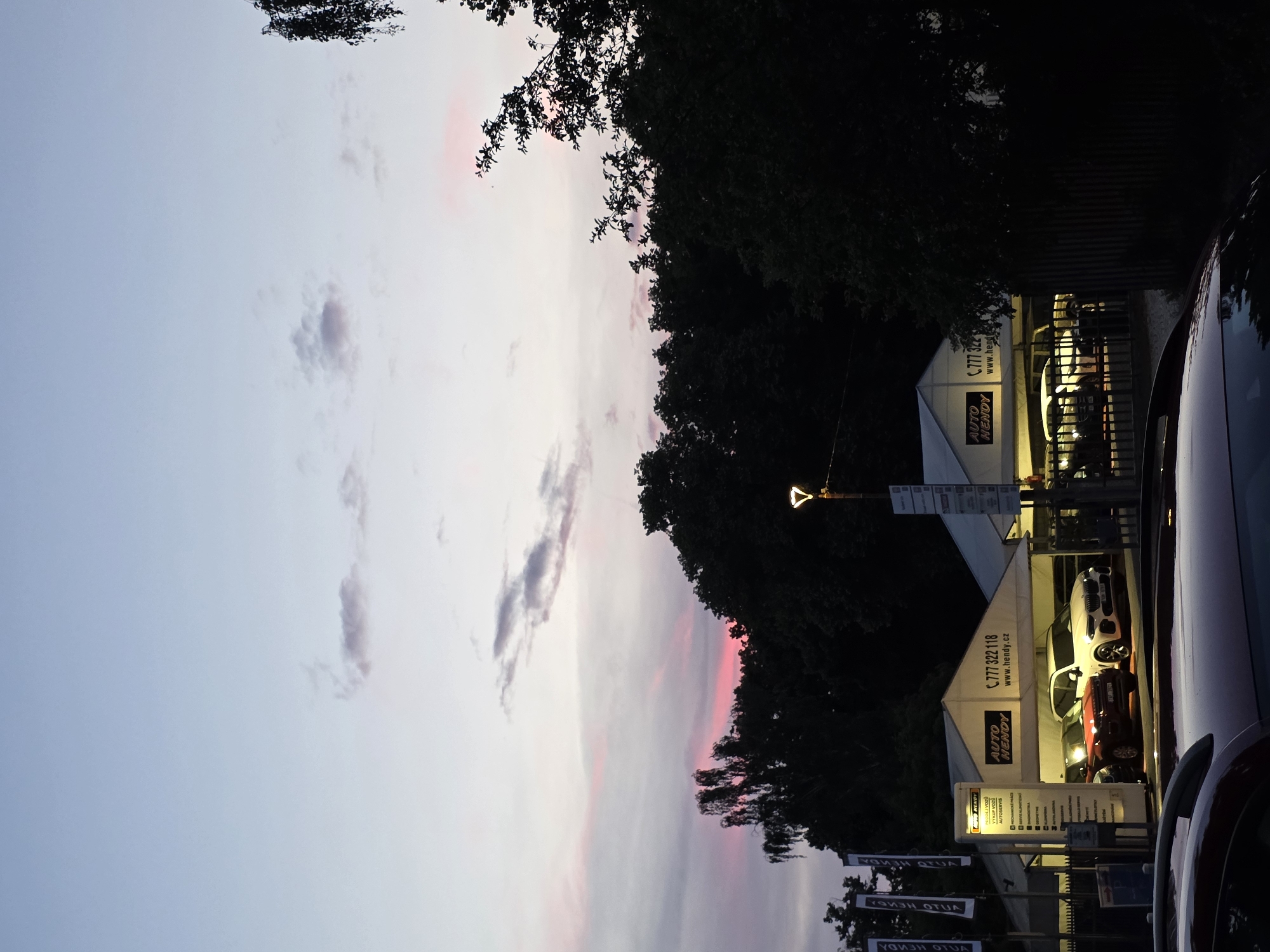 The image shows a car dealership at dusk. Two tents are set up, displaying several cars. One tent has "AUTO HENDY" and a phone number and website printed on it. There are trees and a fence in the foreground and background. The sky is a mix of pink and blue, with some clouds. A street lamp with a unique design is visible on the right side of the image.