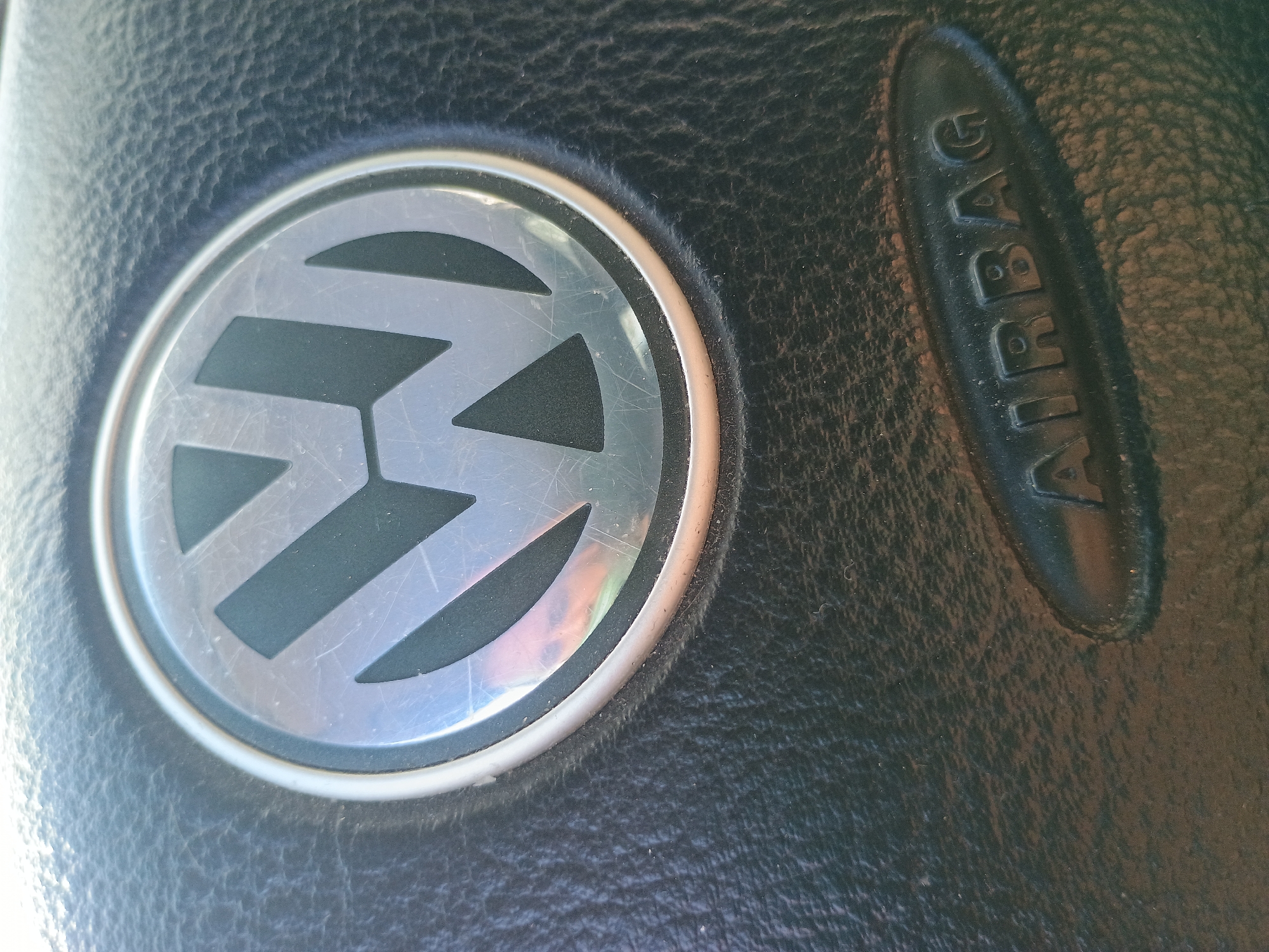 The image shows a close-up of a steering wheel. The prominent features are the Volkswagen logo in the center and the word "AIRBAG" embossed on the right. The material of the steering wheel appears to be textured black leather. Location: Unknown, Unknown.