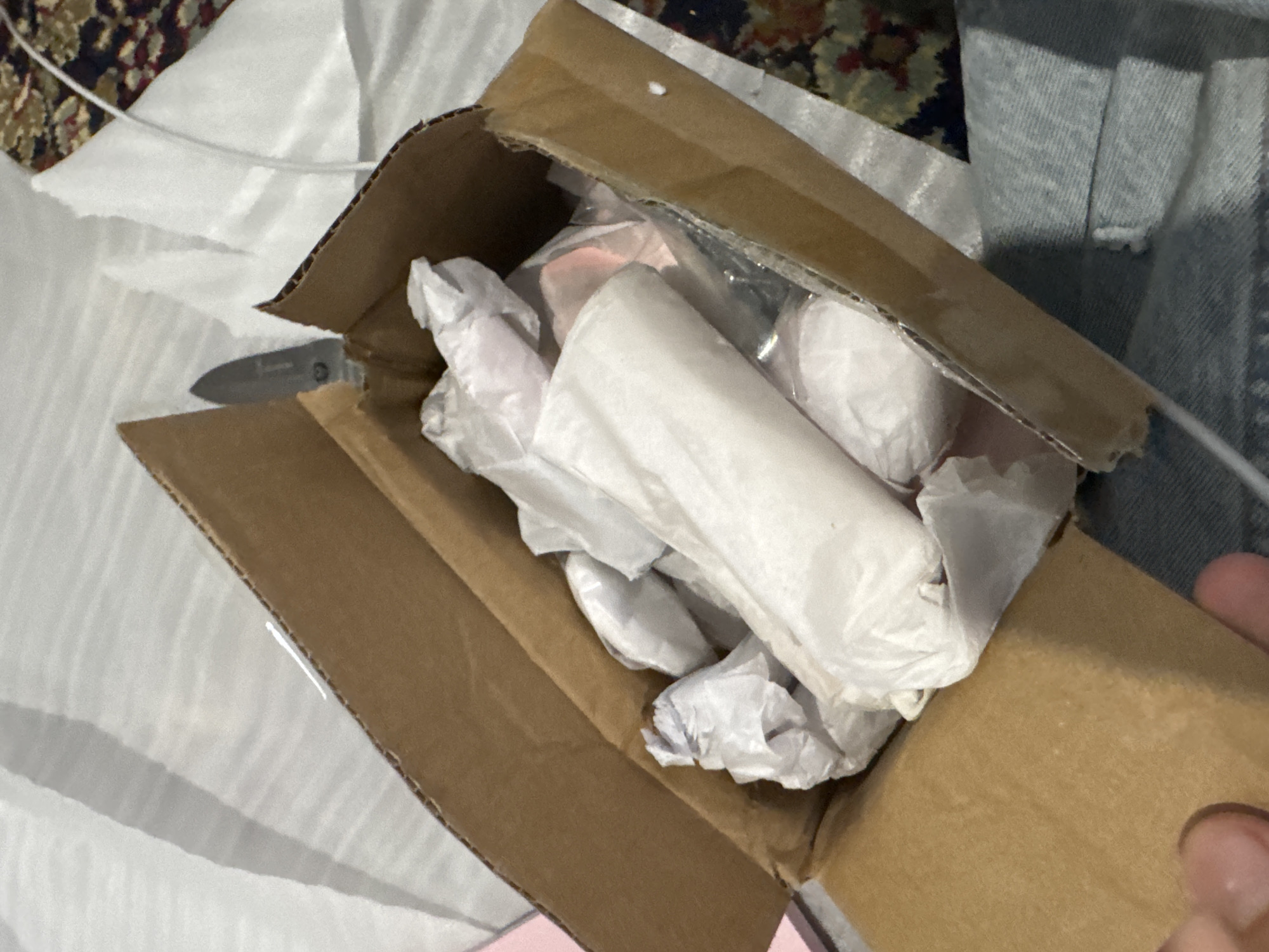 The image shows an opened cardboard box filled with white packing paper. A small utility knife with a silver blade is partially visible, resting on the edge of the box. The box is sitting on a textured surface that appears to be fabric, possibly a rug or carpet with a floral pattern. A portion of what looks like blue denim fabric is also visible at the bottom right of the image. The scene is indoors, with soft lighting, but the exact location within the city of Unknown is not discernible.