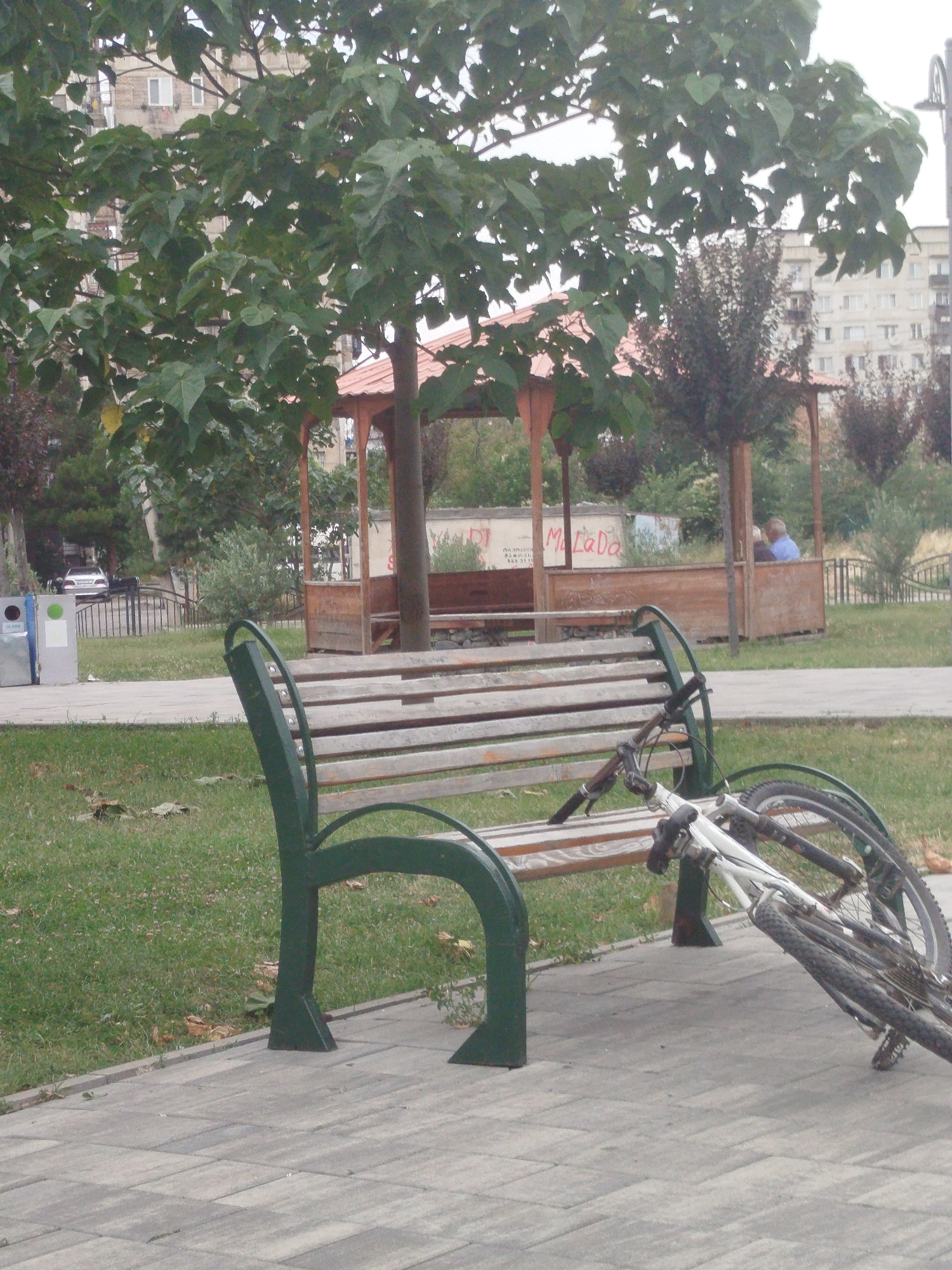 A bicycle is resting on a wooden park bench under a tree, with a wooden gazebo in the background. The scene appears to be in a park in the city of Rustavi, Georgia. The bicycle is white with black accents and has both wheels visible. The bench is made of weathered wood with dark green metal supports. The gazebo has a red-tiled roof and wooden pillars, with people sitting inside. The background shows trees, buildings, and a road with a car. The overall scene suggests a public park setting, possibly during the day.