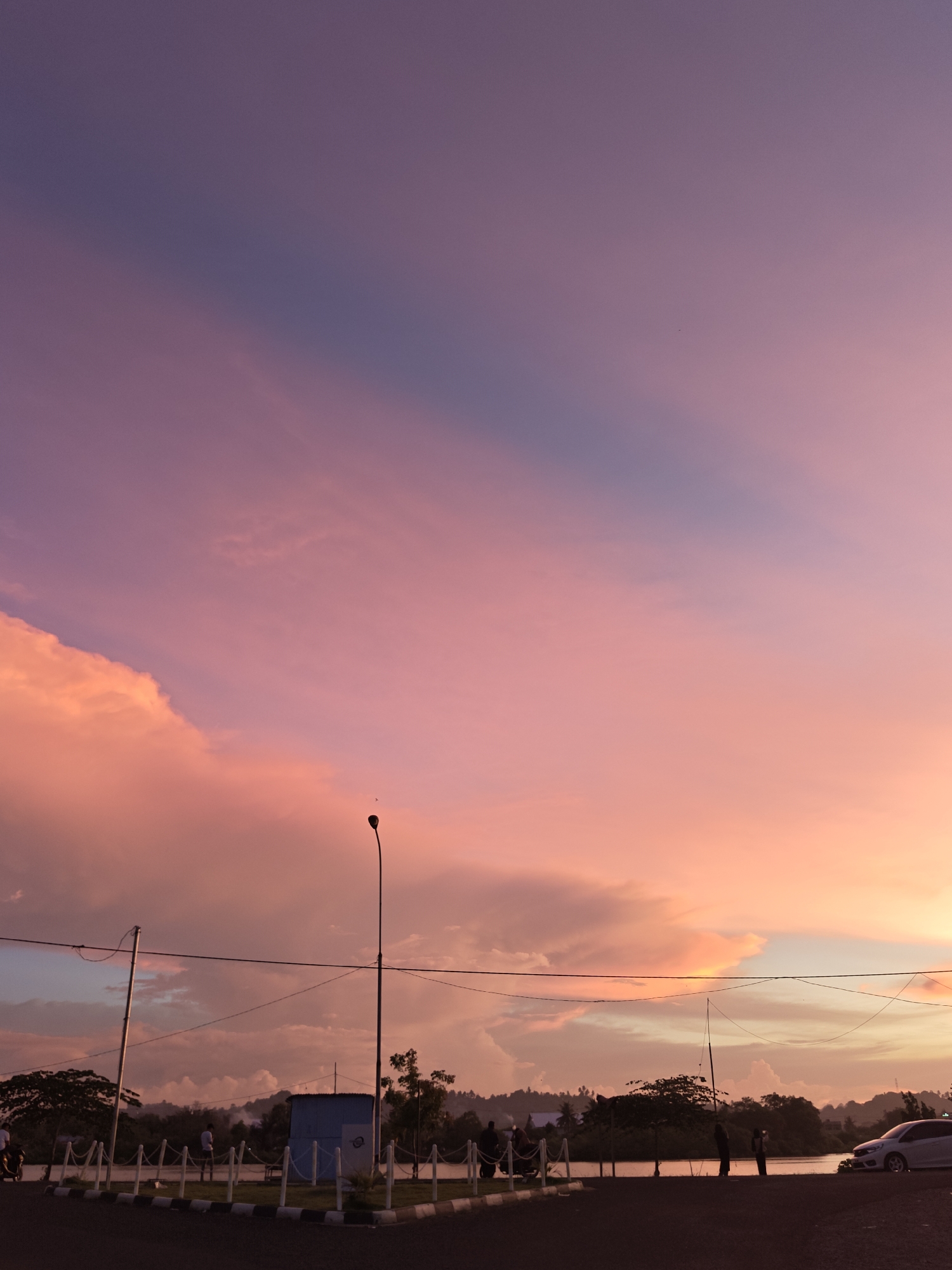 The image depicts a serene sunset scene in Lhokseumawe, Indonesia, likely near the Ujong Blang beach area given the coastal elements. The sky is a vibrant canvas of pink, purple, and orange hues, with soft clouds drifting across. In the foreground, a dark asphalt road curves into the frame. To the left, a white motorcycle with two occupants is visible, suggesting movement or arrival. A paved walkway with a white and black border separates the road from a grassy area. Along this walkway, several individuals are present, some standing and observing the sunset, while others appear to be interacting.
A small, light blue rectangular structure with white trim stands in the middle ground, possibly a utility shed or information booth. A tall, slender lamppost with a single light fixture stands to the right of this structure, its silhouette stark against the colorful sky. Further back, trees and hills form a natural backdrop, with a body of water, likely the sea, visible beyond the grassy area. The silhouette of a white car is parked on the right side of the frame, indicating vehicular presence in the vicinity. The overall atmosphere is tranquil, capturing a moment of natural beauty and quiet activity.