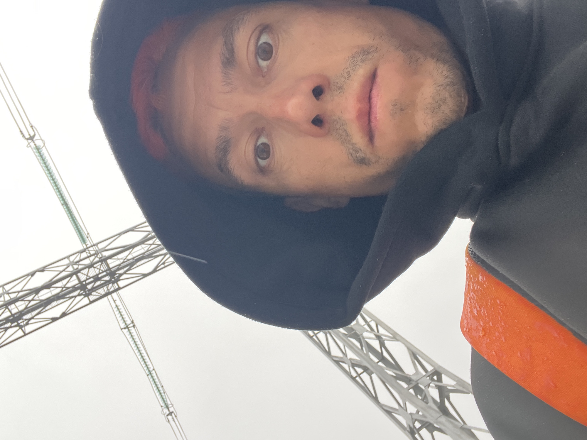 A low-angle, close-up selfie shows a man with light skin, light brown eyes, and reddish hair under a black hood. He has stubble on his chin and a neutral expression. An orange strap is visible on his right. The background shows sections of a power line support structure against a grey sky. The photo appears to have been taken in the city of Chaikovsky, Russia.