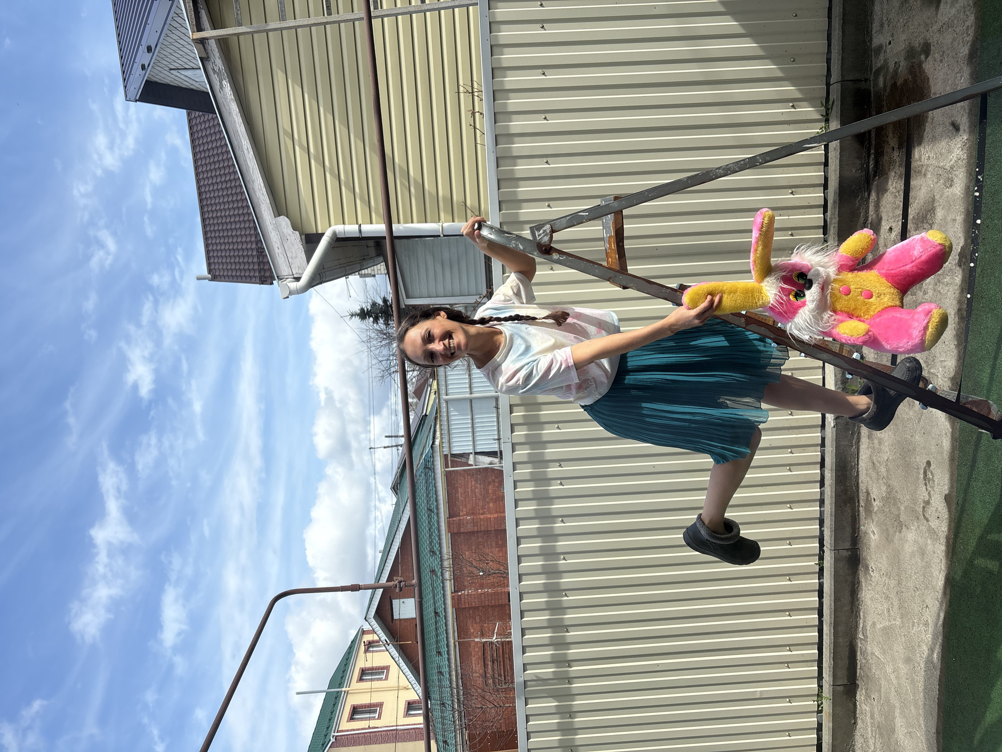 A full-length, rotated image shows a girl standing on a metal structure, possibly a ladder, holding a pink and yellow stuffed rabbit. The girl has long dark hair braided down her back and is wearing a white tie-dye t-shirt and a teal pleated skirt, with black slipper-like shoes. She is smiling and looking towards the camera.
Behind her, a corrugated metal wall serves as a backdrop, and various buildings are visible, including a yellow building with a green roof and a brick building. The sky is blue with scattered clouds. The precise location in Ulyanovsk, Russia cannot be determined from the given details alone. However, it appears to be an outdoor space adjacent to a residential area, possibly a backyard or a side yard.