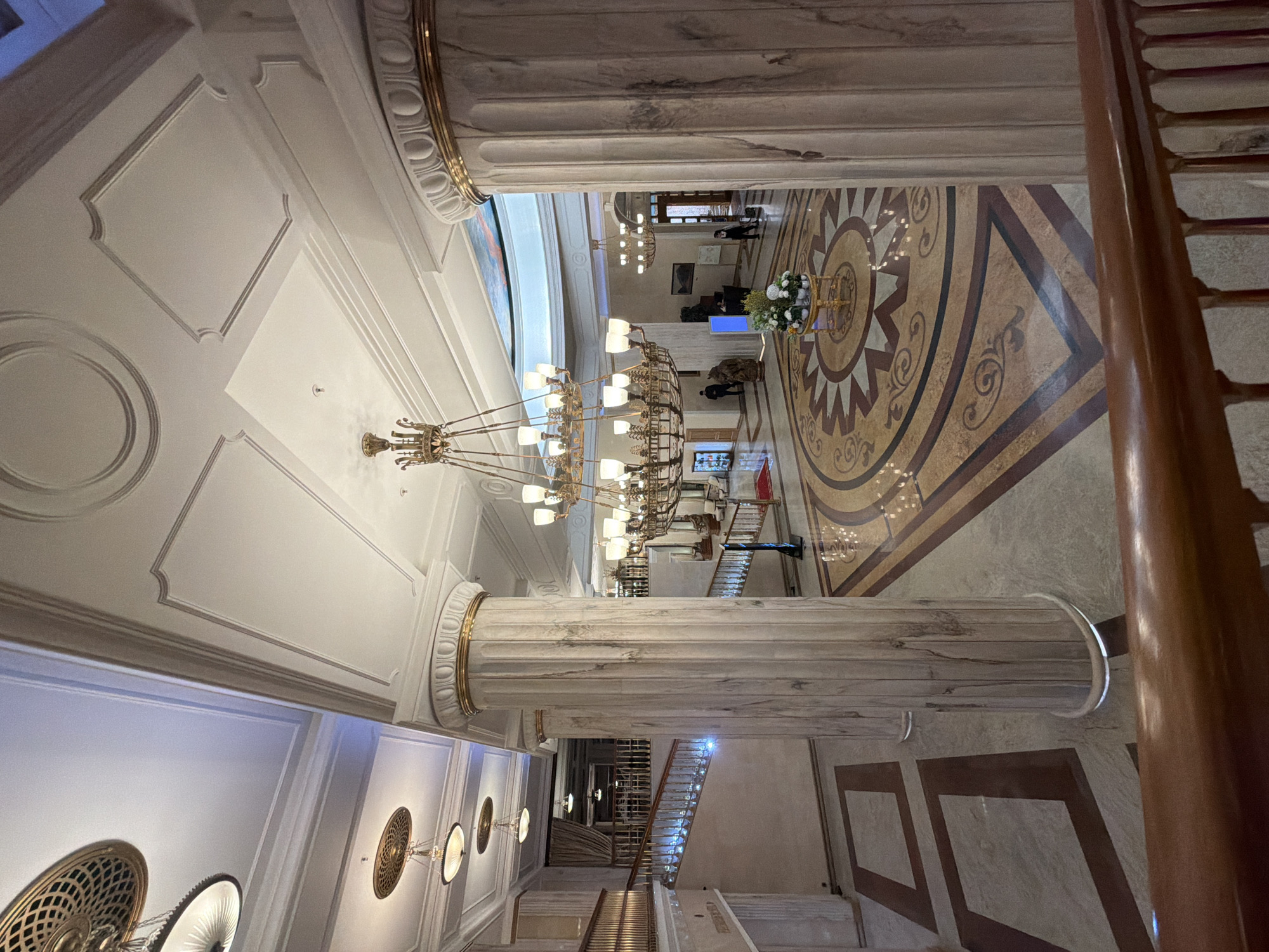 The image captures the opulent interior of a building in Moscow, Russia, likely a historic hotel or government building.
Architectural details include massive marble columns with gold detailing at the top, supporting a high, ornate ceiling with intricate moldings, recessed panels, and decorative lighting fixtures. A grand chandelier hangs prominently in the center of the space.
The floor features a large, patterned mosaic design. Some people are visible in the middle ground of the image. A staircase with a wooden railing is on the right side.