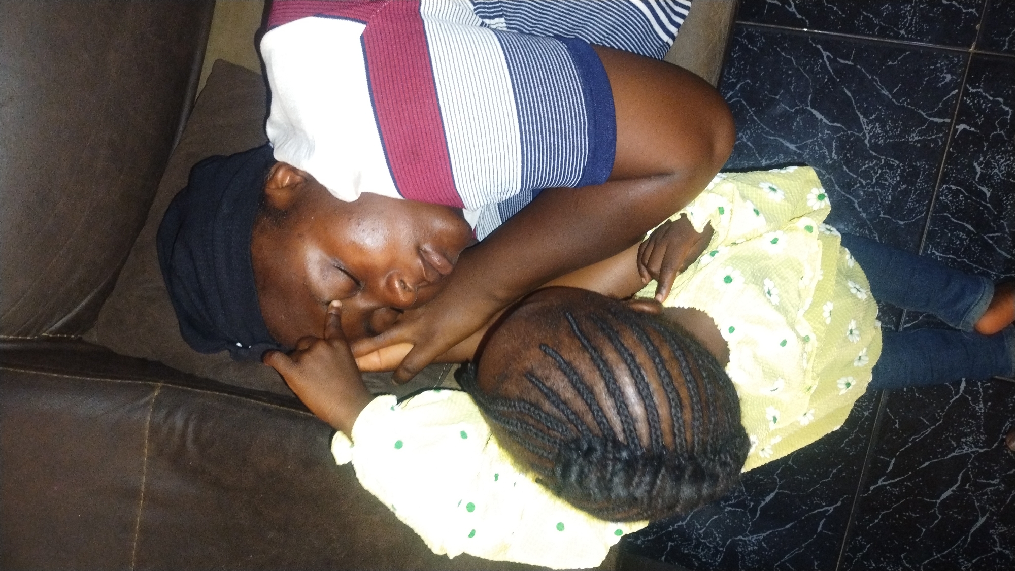 A woman and a child are resting on a brown leather sofa. The woman is wearing a striped shirt and a black head covering. The child, who has braids, is wearing a yellow dress with white floral patterns and blue jeans. The child's hand is touching the woman's face. The location appears to be a residence with a black marble floor. No information is available to confirm the specific location in Jos, Nigeria.