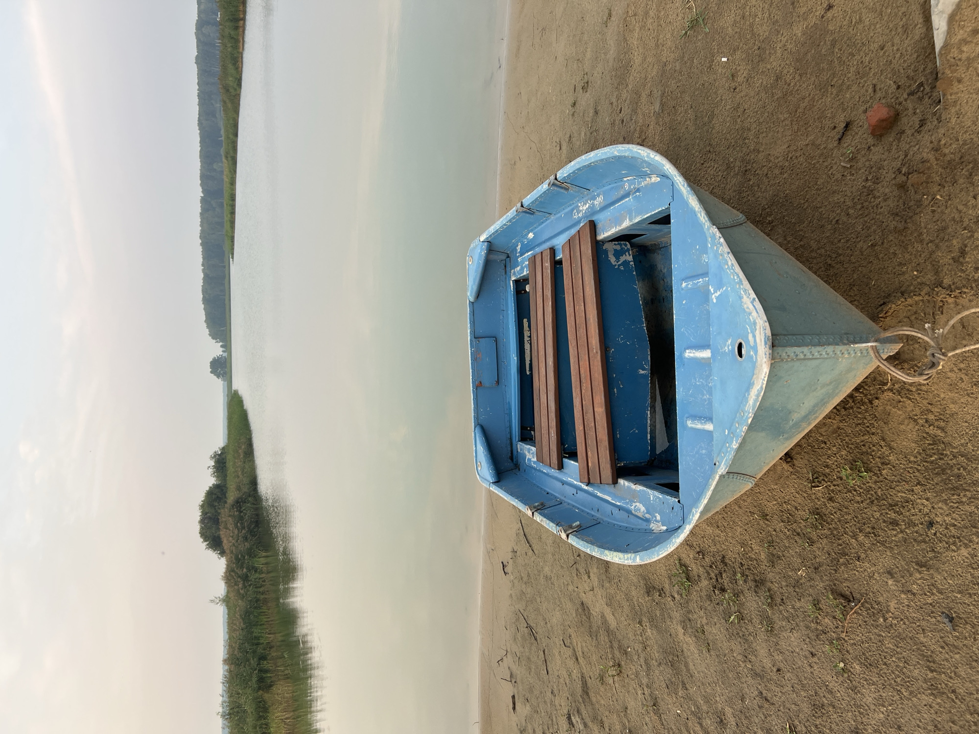 The image depicts a blue metal rowboat resting on a sandy shore next to a calm body of water. The boat is empty and positioned facing the water. Wooden benches are visible inside the boat. The shoreline is characterized by sand and sparse vegetation, with reeds and trees lining the distant banks of the water. The sky is overcast, creating a soft, diffused light. The scene is peaceful and appears to be in a natural, possibly rural, setting, consistent with the location of Novopichugovo, Russia. No people are present in the image.