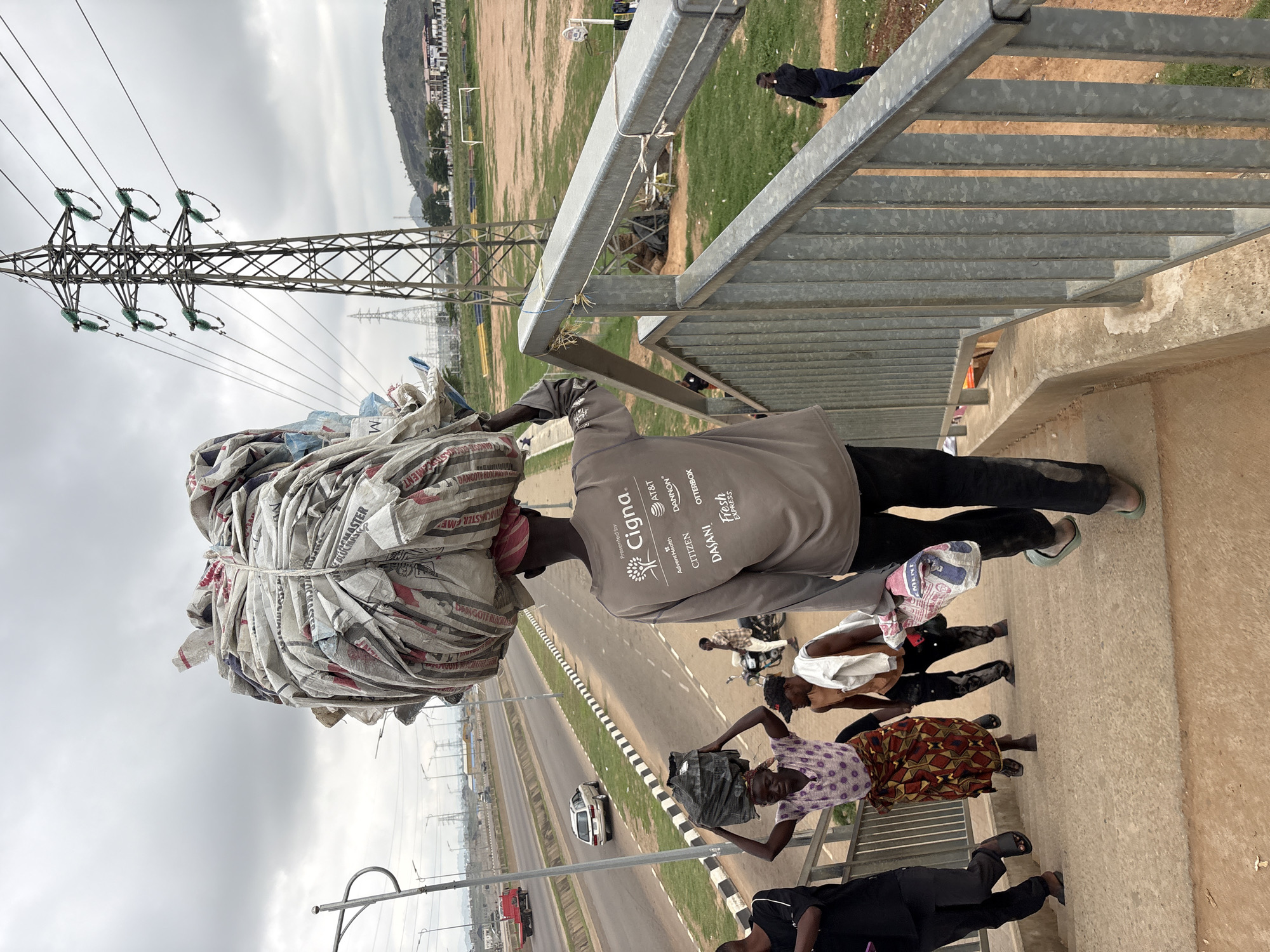In Angwan Sayawa, Nigeria, a group of individuals are depicted on a pedestrian overpass. The central figure, a man, carries a large, tightly bound bundle of what appears to be used cement bags on his head. He is wearing a grey long-sleeved shirt with various logos including "Cigna," "AT&T," and "Citizen." Another man walks behind him carrying a smaller bundle on his head. To the left, a woman wearing a patterned skirt and a purple top carries a dark bag on her head. Two other individuals, a man and a woman, are also visible on the stairs. In the background, a highway with traffic, a power pylon, and a grassy field with goalposts are observed. The sky is overcast.