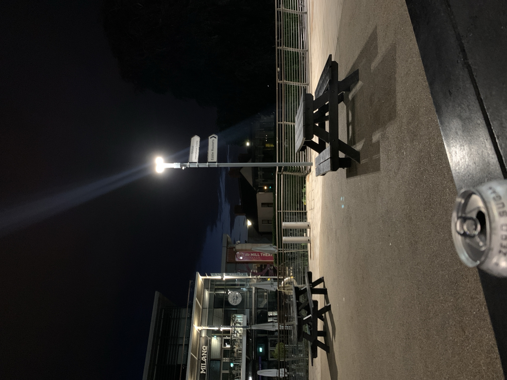 The image depicts an outdoor scene in Dublin 16, Ireland at night. A bright street lamp illuminates an area with restaurants, including one named "Milano" and signs pointing to "dlr Mill Theatre". There are metal benches and tables present, suggesting an outdoor dining area. An empty, crushed soda can lies discarded on the ground. People are vaguely visible in the illuminated restaurant. The overall environment suggests a commercial district at night.