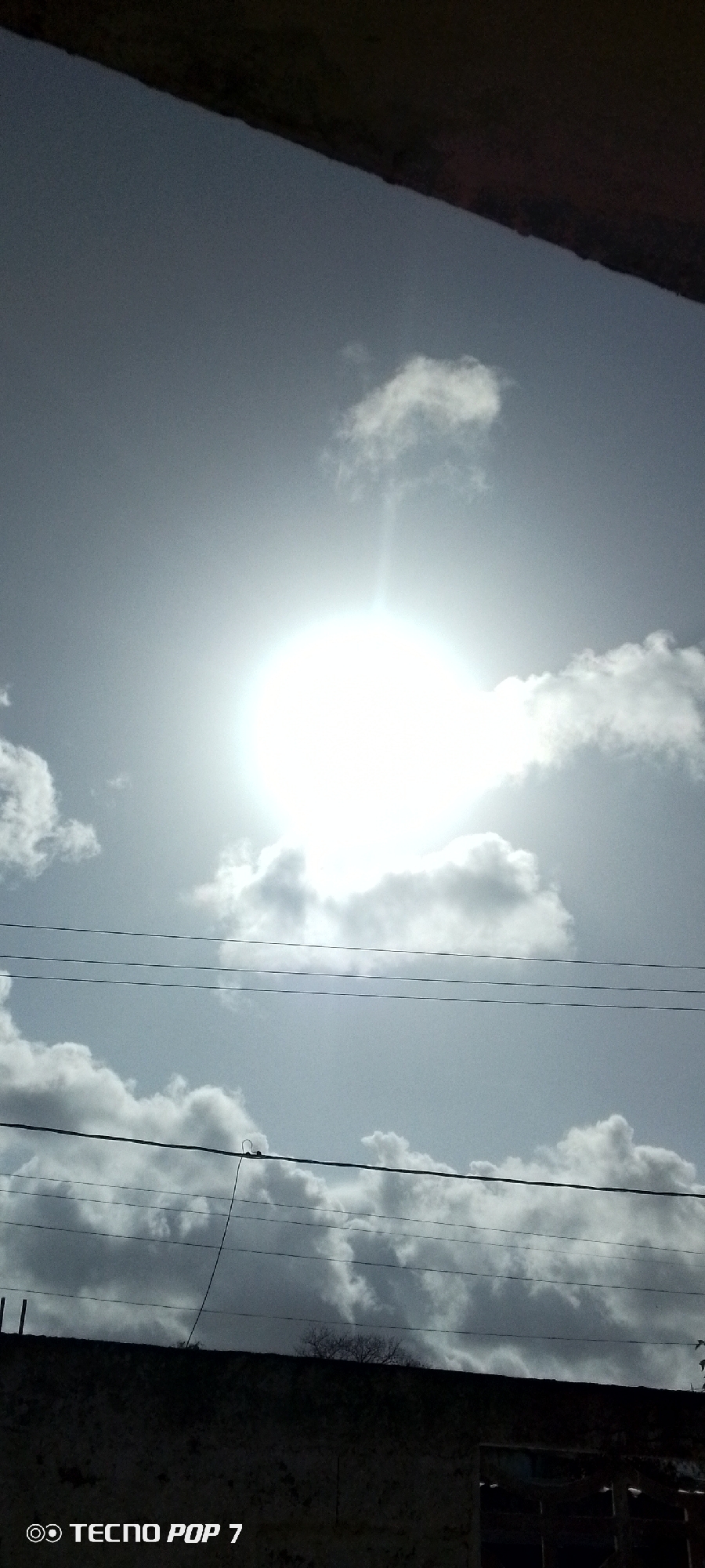 The image shows a daytime sky with bright sunlight obscured by thin clouds. Several utility lines stretch horizontally across the frame. Below the lines, a dark wall with some vegetation along the top is visible. The photo was taken in Altagracia, Venezuela, as indicated by the camera settings.