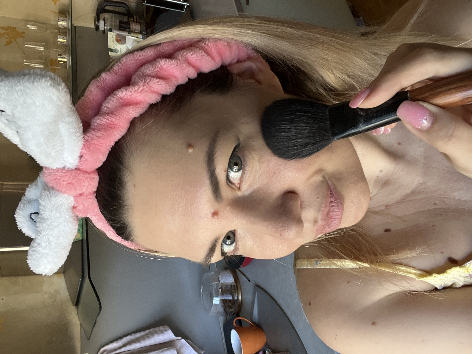 A young Caucasian woman is applying makeup in a kitchen. She has long blonde hair and is wearing a pink headband with white bunny ears. She is holding a black makeup brush with a wooden handle and has pink, glittery nail polish on her fingers. She is wearing a yellow, floral print top. The kitchen appears to be in an apartment in Sochi, Russia, with cabinets and shelves visible in the background. There is an orange mug on the counter.