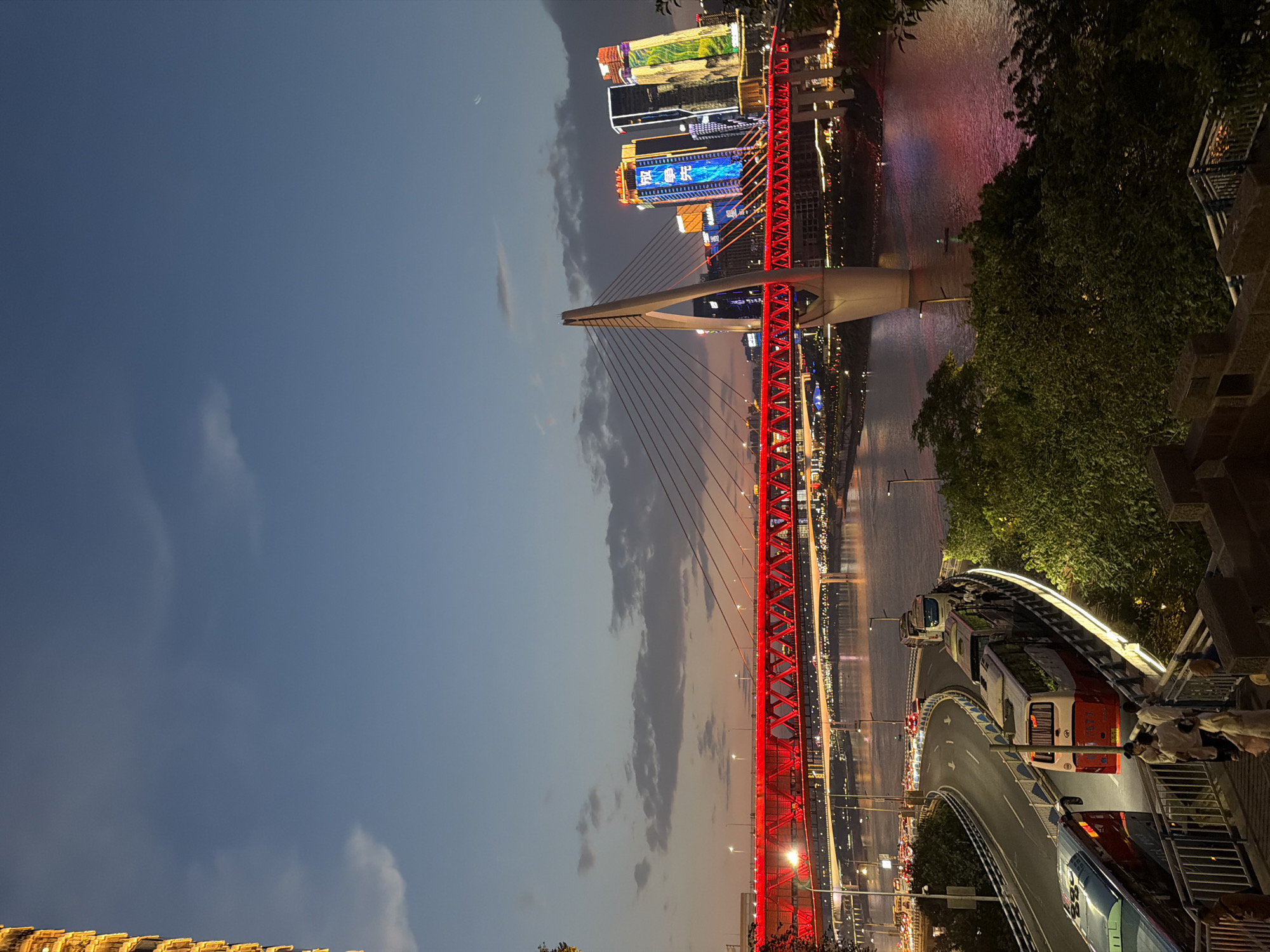 This image captures a twilight view of the Hongyadong Bridge in Yuzhong District, Chongqing, China. The bridge, illuminated with vibrant red lights, spans across a wide river. Tall, modern buildings adorned with colorful neon lights rise in the background. A road with flowing traffic, including several buses, curves alongside the river. In the foreground, a set of stairs leads down from a vantage point offering a view of the scene. The sky is a deep blue with scattered clouds.