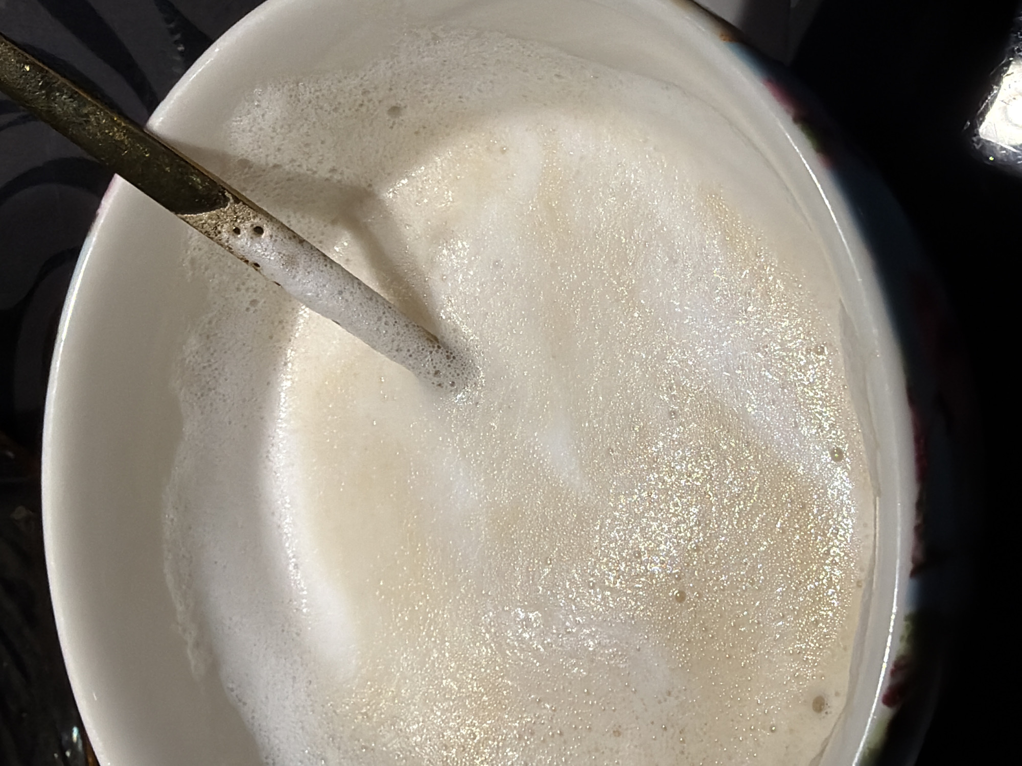 The image depicts a close-up, overhead shot of a white bowl filled with a frothy, light-colored liquid, likely a beverage. A metallic straw or spoon is partially submerged in the liquid, positioned on the left side of the frame. The liquid exhibits varying textures, with some areas appearing smoother and others more textured with visible bubbles. The bowl's rim is visible along the edges of the frame, revealing a slight decorative pattern. The background is dark and out of focus, possibly indicating a table or countertop surface. The image was taken in Rossiyskiy, Russia, as per the user's prompt.
