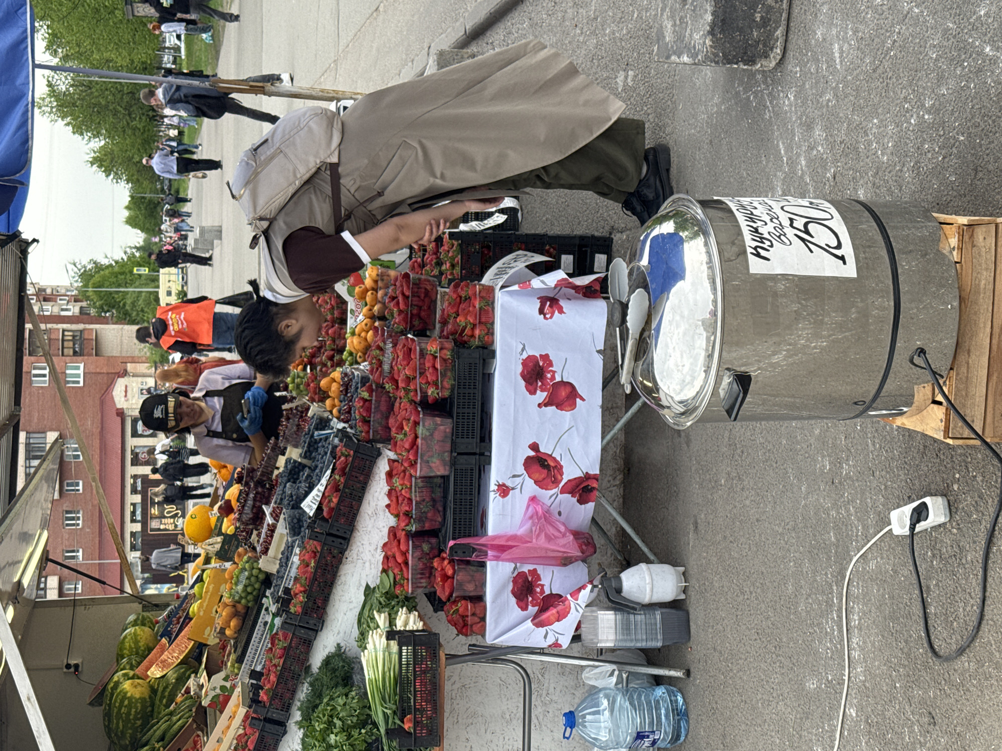 The image shows a street market in Saint Petersburg, Russia. A woman wearing a tan jacket and dark pants is interacting with a customer at a fruit stand. The stand displays a variety of fruits including strawberries, oranges, watermelons, and blueberries in black plastic containers. A white tablecloth with a poppy design covers a table next to the fruit. Beside the table is a large metal container labeled "150", possibly indicating the price. Other people are walking in the background along a paved path. Buildings and trees are also visible in the background. A multi-outlet adapter and several power cords are present.