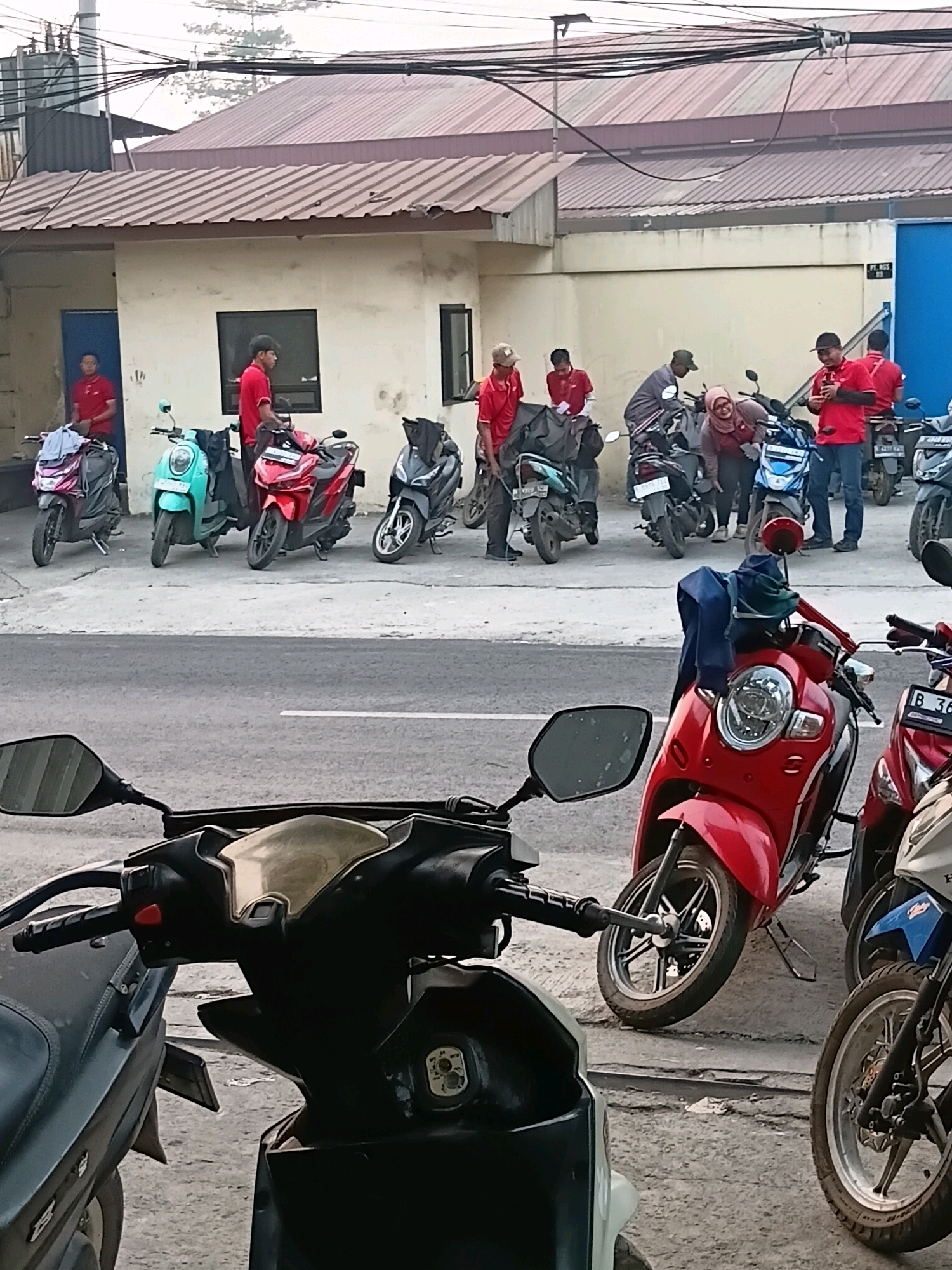 This image depicts a group of delivery personnel gathered with their scooters outside a building in Bekasi, Indonesia. The scene is a street-level view, with several motor scooters parked in a row on the paved ground in front of a single-story building. The building features a light-colored facade, a brown corrugated metal roof, and a blue doorway on the left.
Several individuals, likely delivery workers, are present. Most are wearing red shirts, suggesting a uniform. Some are standing near their scooters, while others are engaged in conversation or preparing their deliveries. One woman in a red headscarf is interacting with a scooter. The scooters are various colors, including red, turquoise, gray, and blue, with some carrying large black delivery bags.
In the foreground, the handlebars and mirrors of several more scooters are visible, indicating a dense parking area. The environment suggests an urban or semi-urban setting, with overhead electrical wires and a glimpse of trees in the background. The lighting indicates it is daytime, possibly late afternoon, with a slightly overcast sky. The overall impression is one of an active delivery hub or staging area.