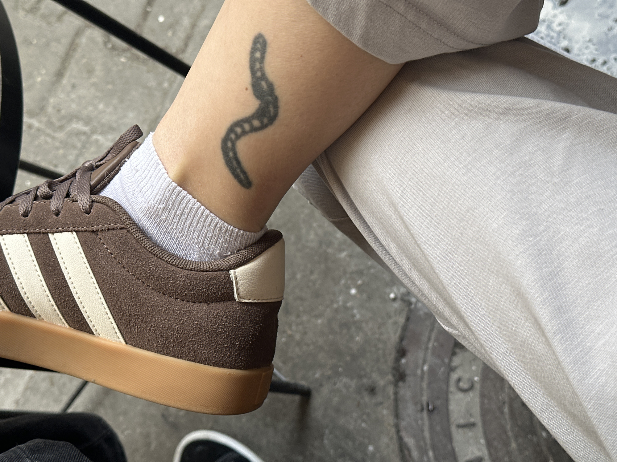 The image provides a close-up, high-angle perspective of a person's lower leg and foot, resting in what appears to be an outdoor setting in Novi Sad, Serbia.
The central subject is a brown suede sneaker, featuring three cream-colored stripes on the side, brown laces, a matching cream heel tab, and a distinctive tan/gum rubber sole. The shoe is worn over a white, ribbed ankle sock. Above the sock, on the person's lower calf/ankle, a black tattoo is visible, depicting a stylized, segmented, and wavy shape, reminiscent of a worm or centipede. The person is also wearing light-colored (possibly linen) trousers, a portion of which is visible draped over their upper leg.
The scene is set on a grey, paved surface, likely concrete or flagstones. In the background, to the lower right, a circular metal manhole cover or utility grate is partially visible. Fragments of embossed text can be discerned on it, including "I.C." and "L," potentially part of a longer word such as "KANALIZACIJA" (sewerage) common on such covers in Serbia. A dark, thin metal object, possibly a furniture leg, is partially seen on the left side of the frame. The lighting suggests it is daytime, and the overall atmosphere is casual and relaxed.