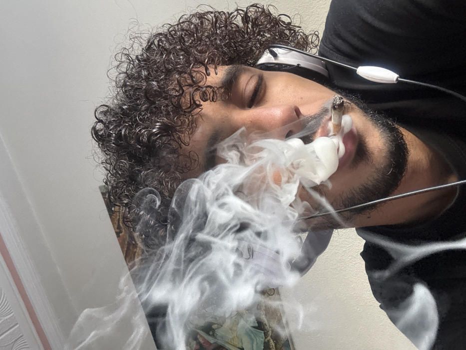 This close-up image depicts a male individual in Tangier, Morocco. He has dark, curly hair, light skin, and a beard/stubble, with his eyes closed. A hand-rolled cigarette is held between his lips, releasing a significant volume of white smoke that billows upwards, partially obscuring his face. He wears white over-ear headphones, connected by a visible white cord with an inline control unit, and a black t-shirt. The background reveals a light-colored wall, with a framed artwork or tapestry featuring blue, green, and brown tones partially visible in the upper left. A white ceiling with decorative molding is present above the artwork.