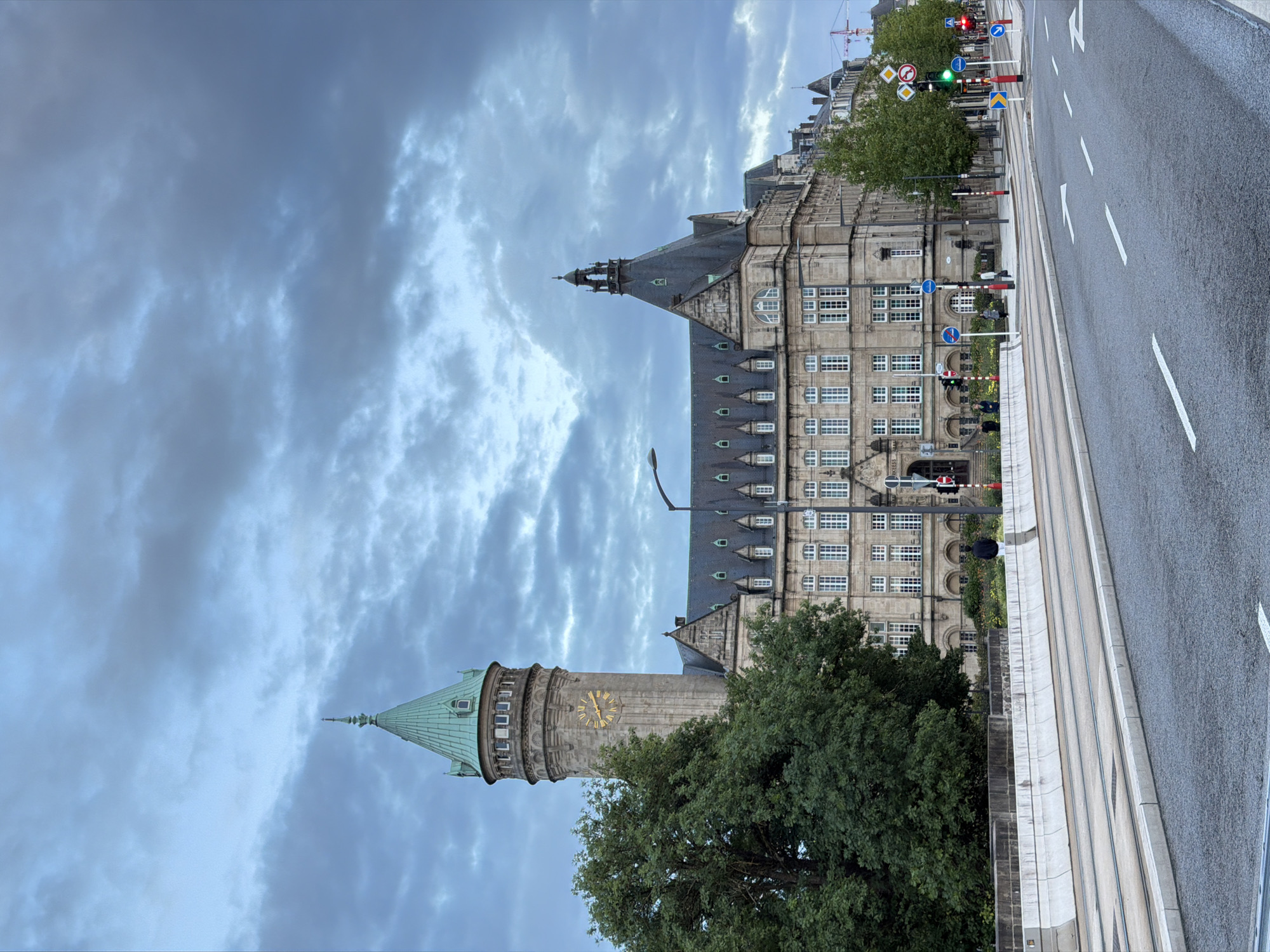 The image depicts the BCEE Building, also known as Spuerkeess, in Luxembourg City, Luxembourg. The imposing stone structure features a prominent clock tower with a green conical roof and a secondary, smaller spire. The main building exhibits classic architectural elements, including numerous windows and dormers. To the left of the tower, a large, mature tree with dense green foliage is visible. In the foreground, a street with tram tracks runs parallel to a wide sidewalk or pedestrian area. Traffic lights and road signage are present along the street. Several individuals are walking or standing on the pedestrian area. The sky is overcast with dramatic clouds, suggesting a moody or stormy atmosphere. The overall scene is one of urban architecture set against a natural backdrop and a dynamic sky.