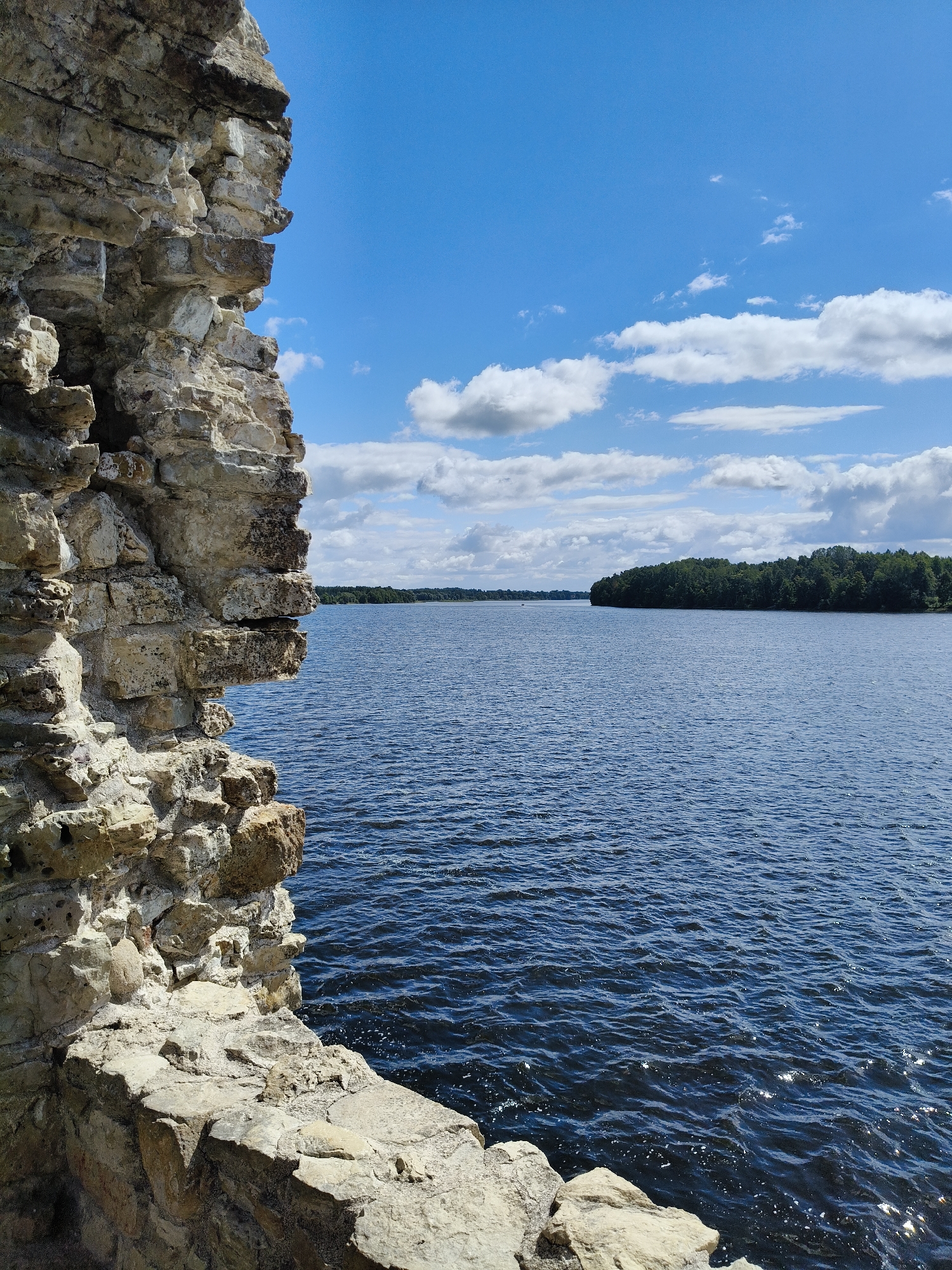 The image depicts the ruins of Koknese Castle in Koknese, Latvia, as seen from a partially collapsed stone wall. The wall, made of rough-hewn light-colored stones, occupies the left side of the frame, with its textured surface and varied rock formations creating a strong foreground element. Beyond the wall, a large body of dark blue water stretches towards the horizon, its surface rippled by a gentle breeze and reflecting the sunlight. The distant shore is lined with a dense green forest, and further in the distance, the horizon is a soft blend of water and land under a bright blue sky dotted with white, fluffy clouds. The scene is tranquil and evokes a sense of historical grandeur mixed with natural beauty. No people are visible in the image.
